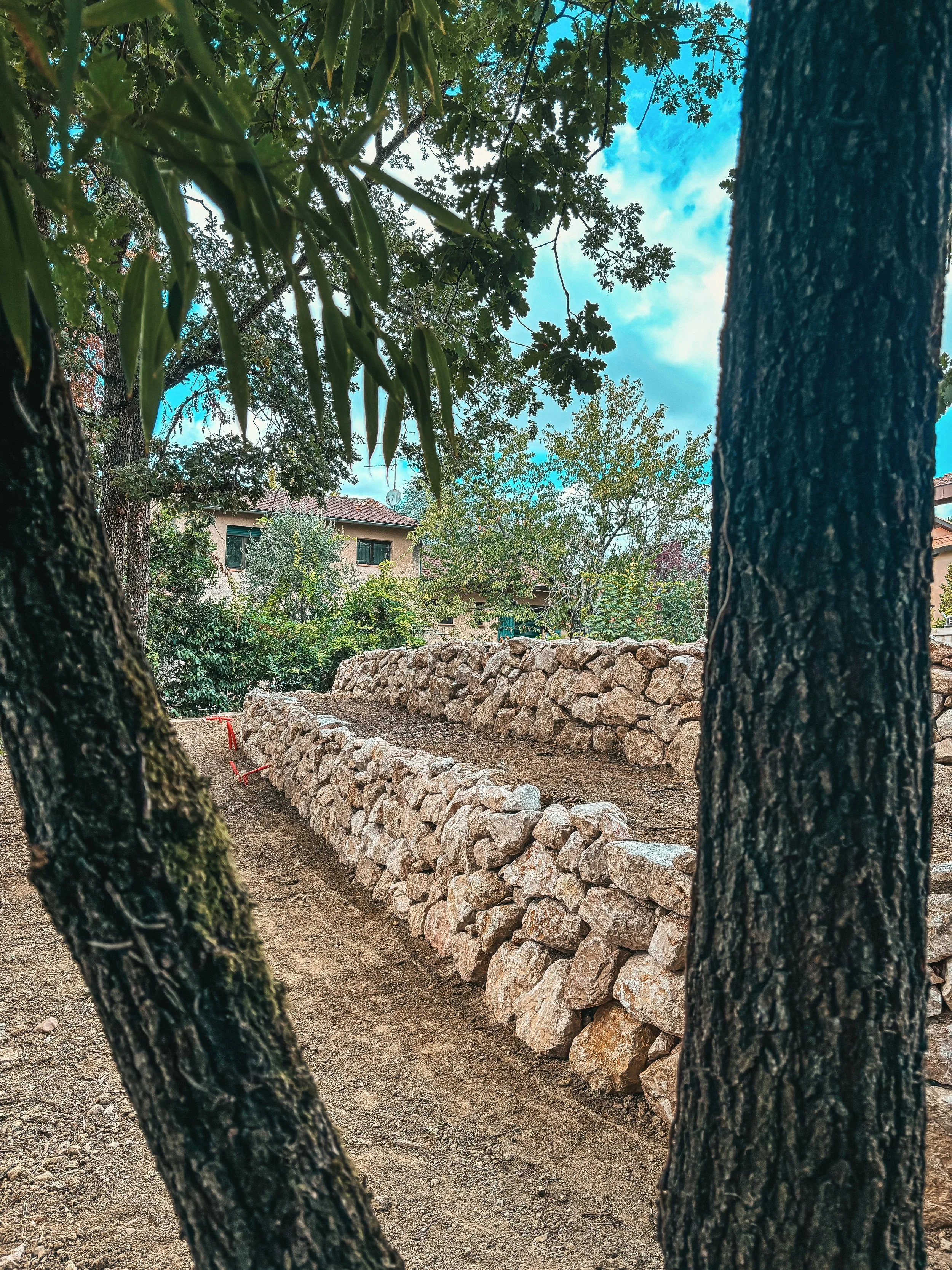 Une scene de jardin avec deux arbres en premier plan, une haie en pierre, une maison en arrière-plan, et un ciel partiellement nuageux.