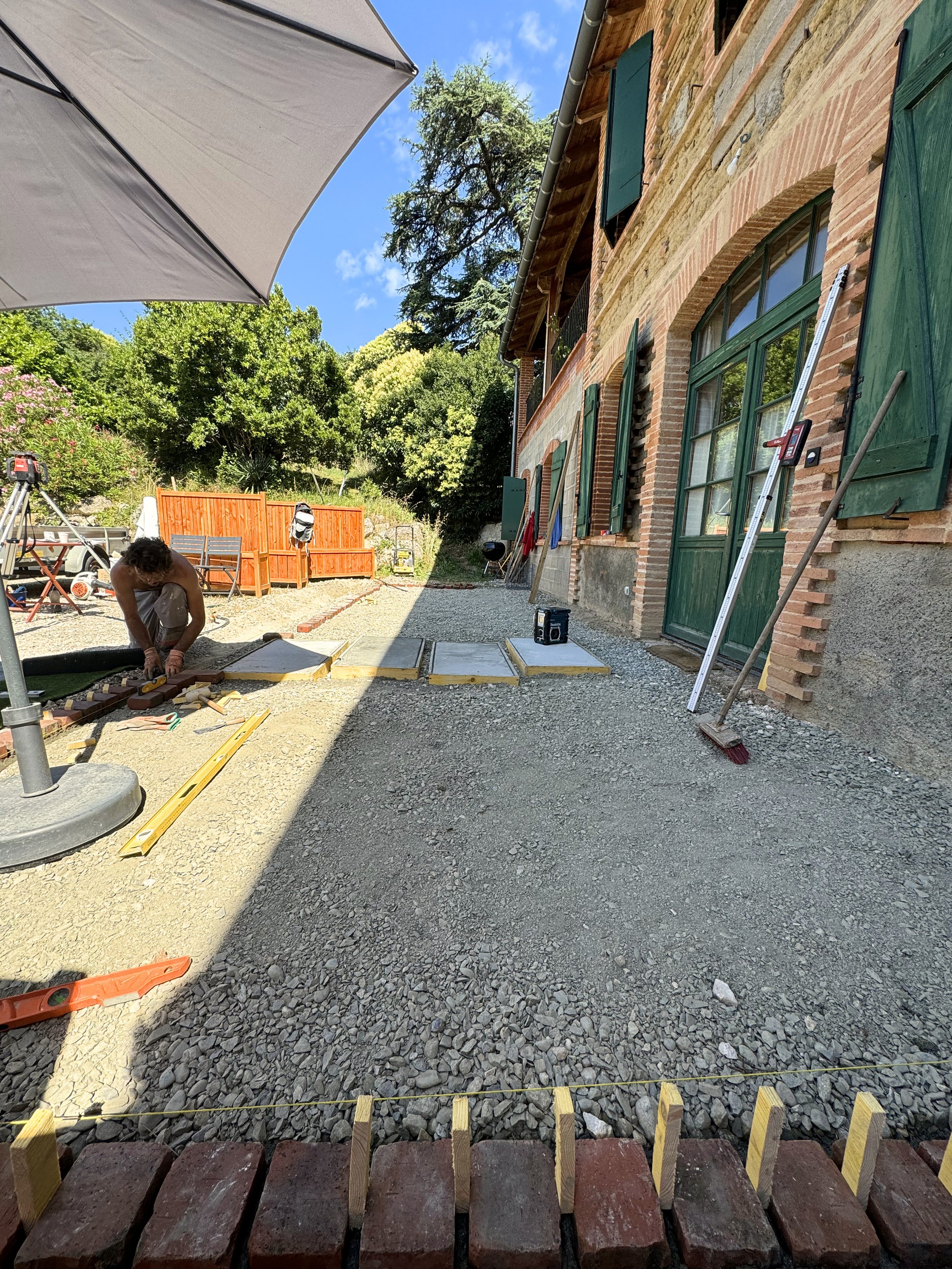 Travail de construction ou de rénovation en cours dans la cour d'une maison en pierre avec volets verts, outils et matériaux de chantier, et un homme travaillant au sol, sous un parasol avec un ciel bleu et des arbres en arrière-plan.