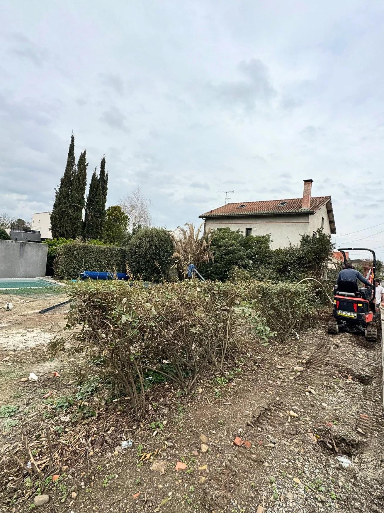 Un jardin en train d'être nettoyé, avec des buissons et des arbres, une maison en arrière-plan, et une personne utilisant une petite machine pour couper des branches.