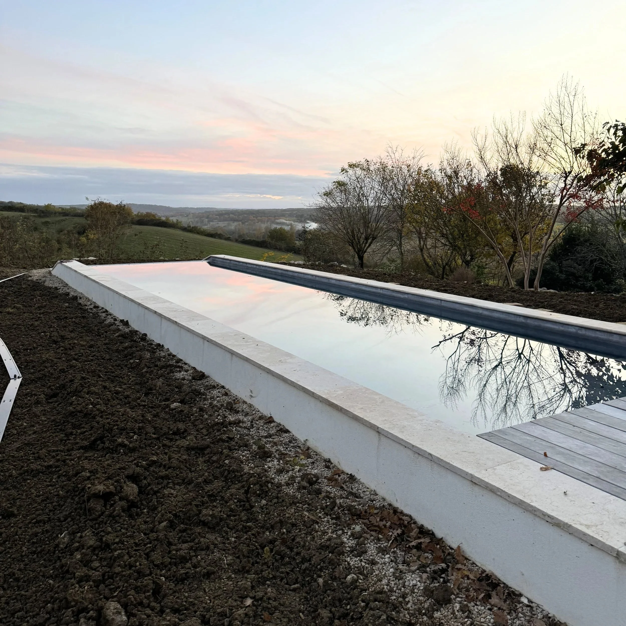 Vue d'ensemble d'un paysage rural avec un ciel nuageux à l'aube, des arbres aux feuilles automnales et un bassin d'eau en construction dans un terrain en terre.