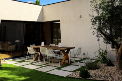 Une terrasse extérieure avec une table en bois, des chaises blanches, et un jardin avec des arbustes et un arbre à proximité, face à une maison moderne.