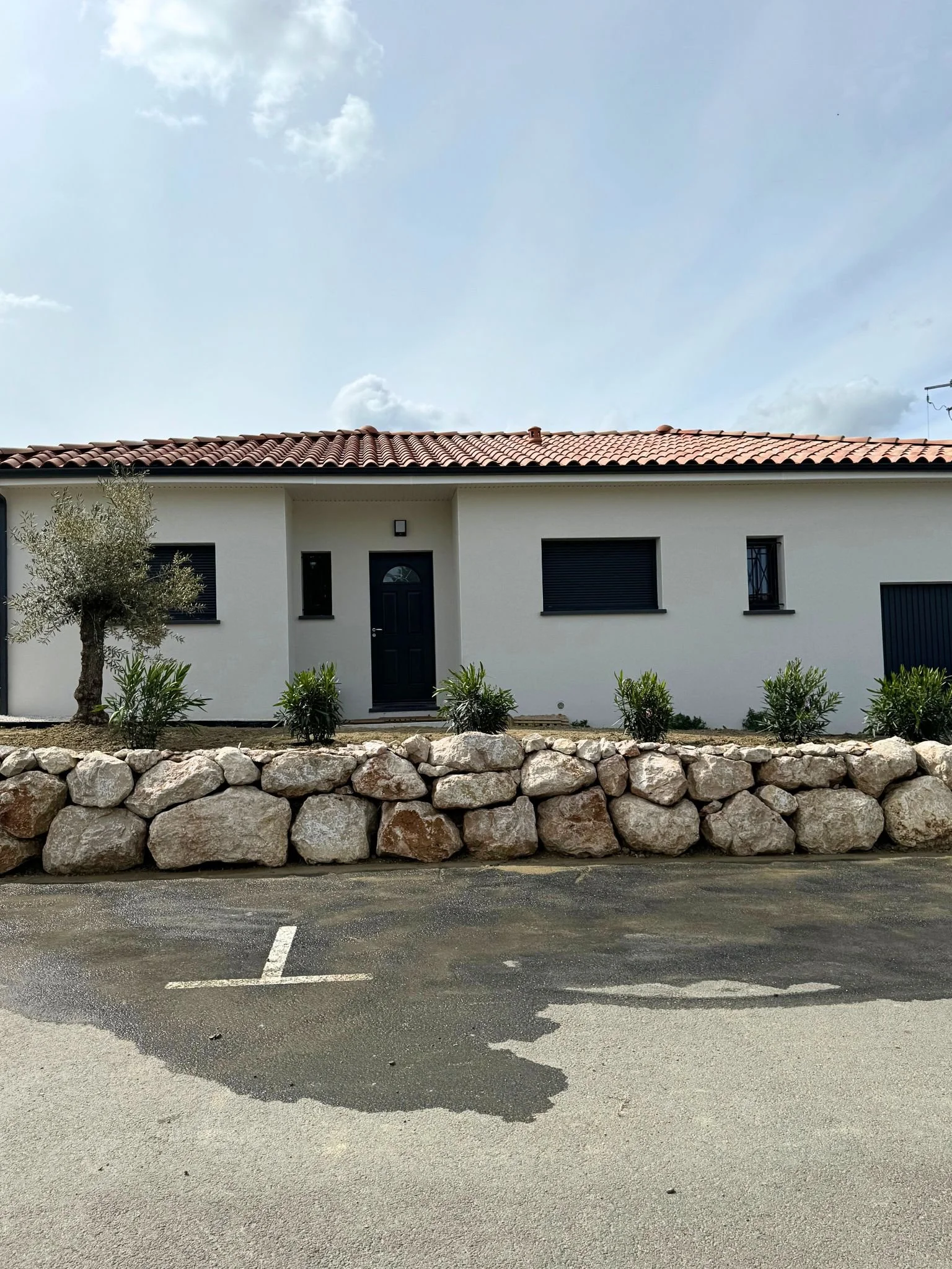 Une maison blanche avec un toit en tuiles rouges, des fenêtres avec volets noirs, un arbre et des buissons devant. Il y a un mur en pierre devant la maison, et une place de parking vide avec des lignes blanches au sol.