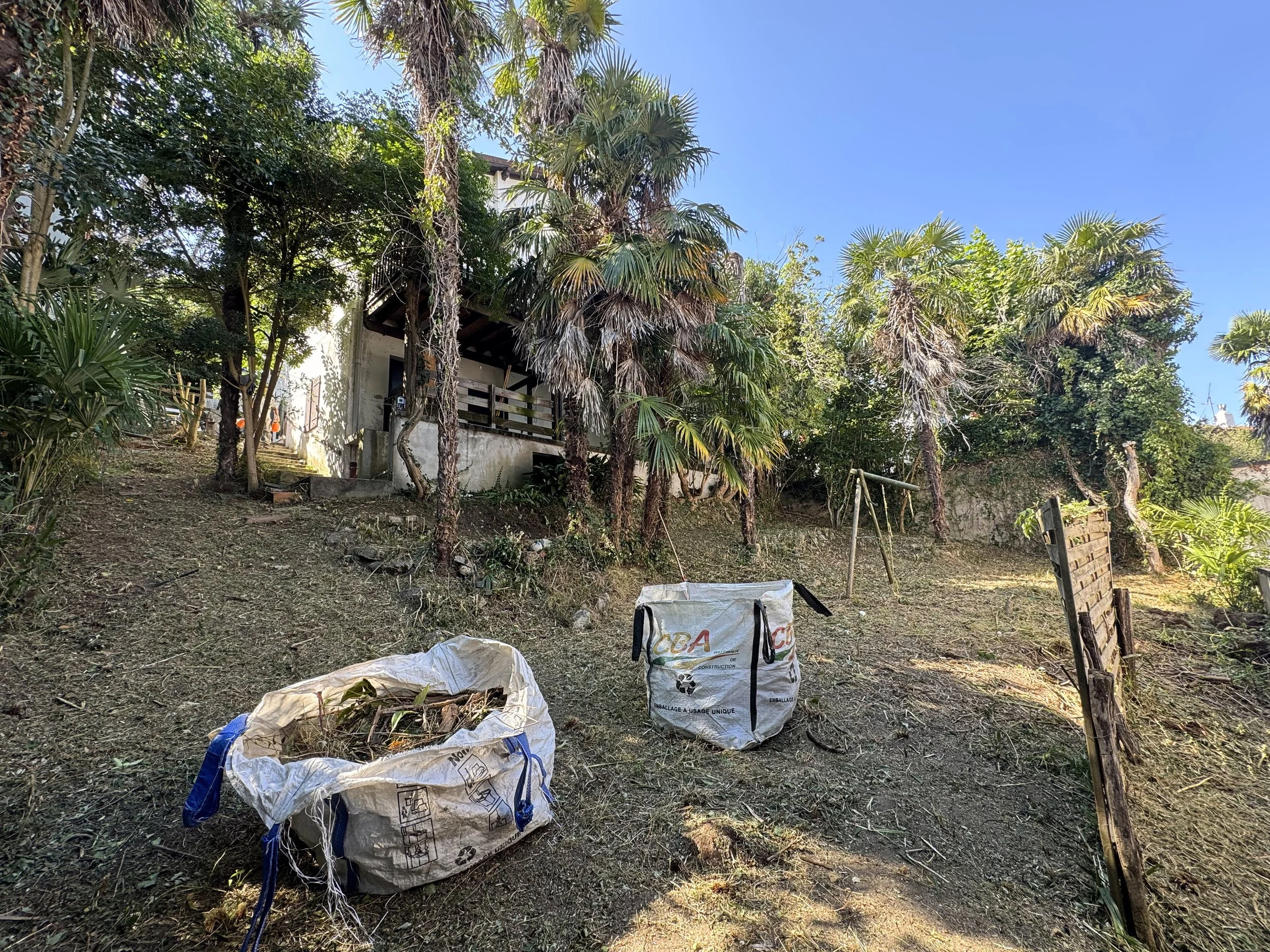 Un jardin avec de nombreux arbres et une maison partiellement visible en arrière-plan, des sacs de déchets ou de matériaux de construction au premier plan.