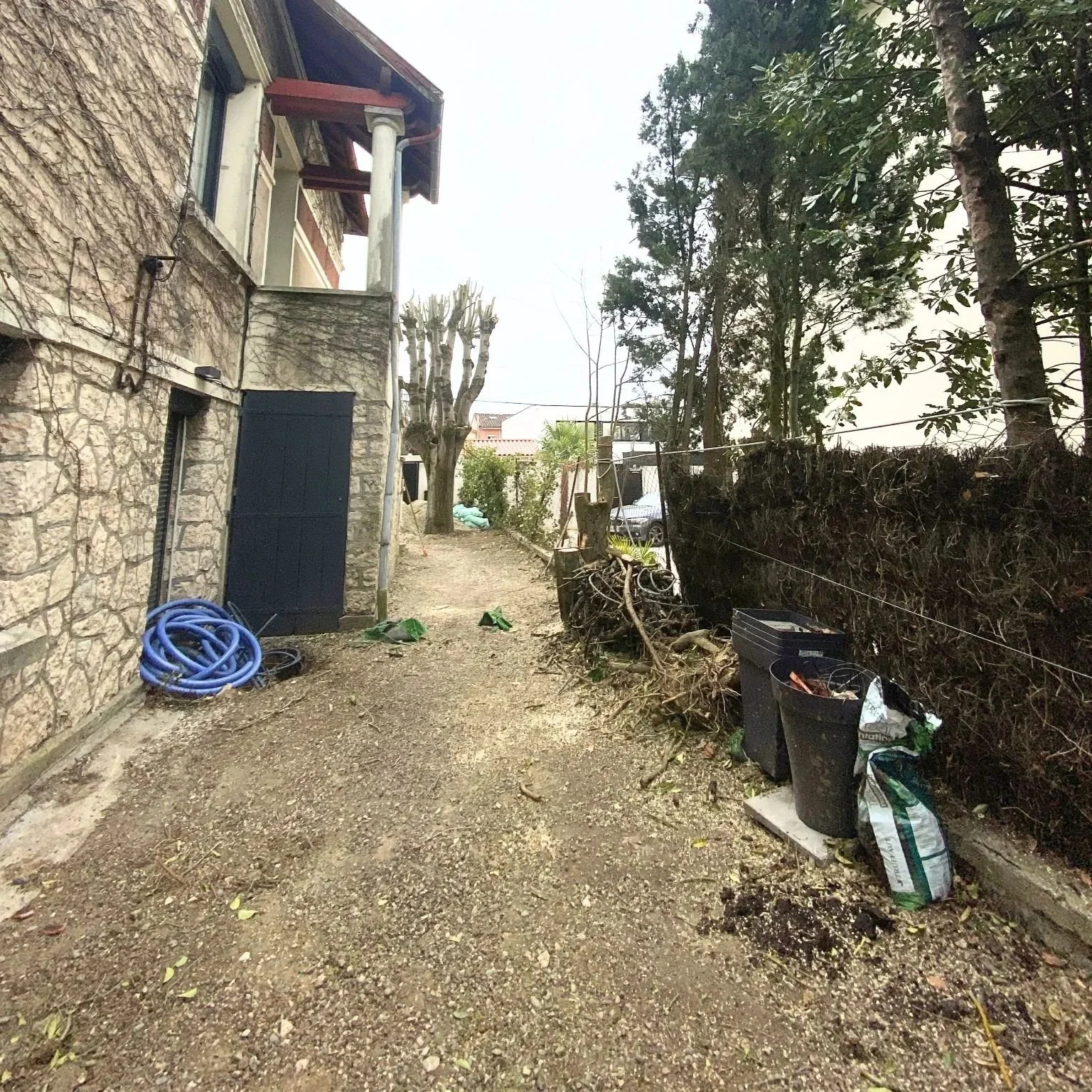 Une cour extérieure bordée d'un mur en pierre, avec des plantes, des déchets, et des outils de jardinage, sous un ciel nuageux.