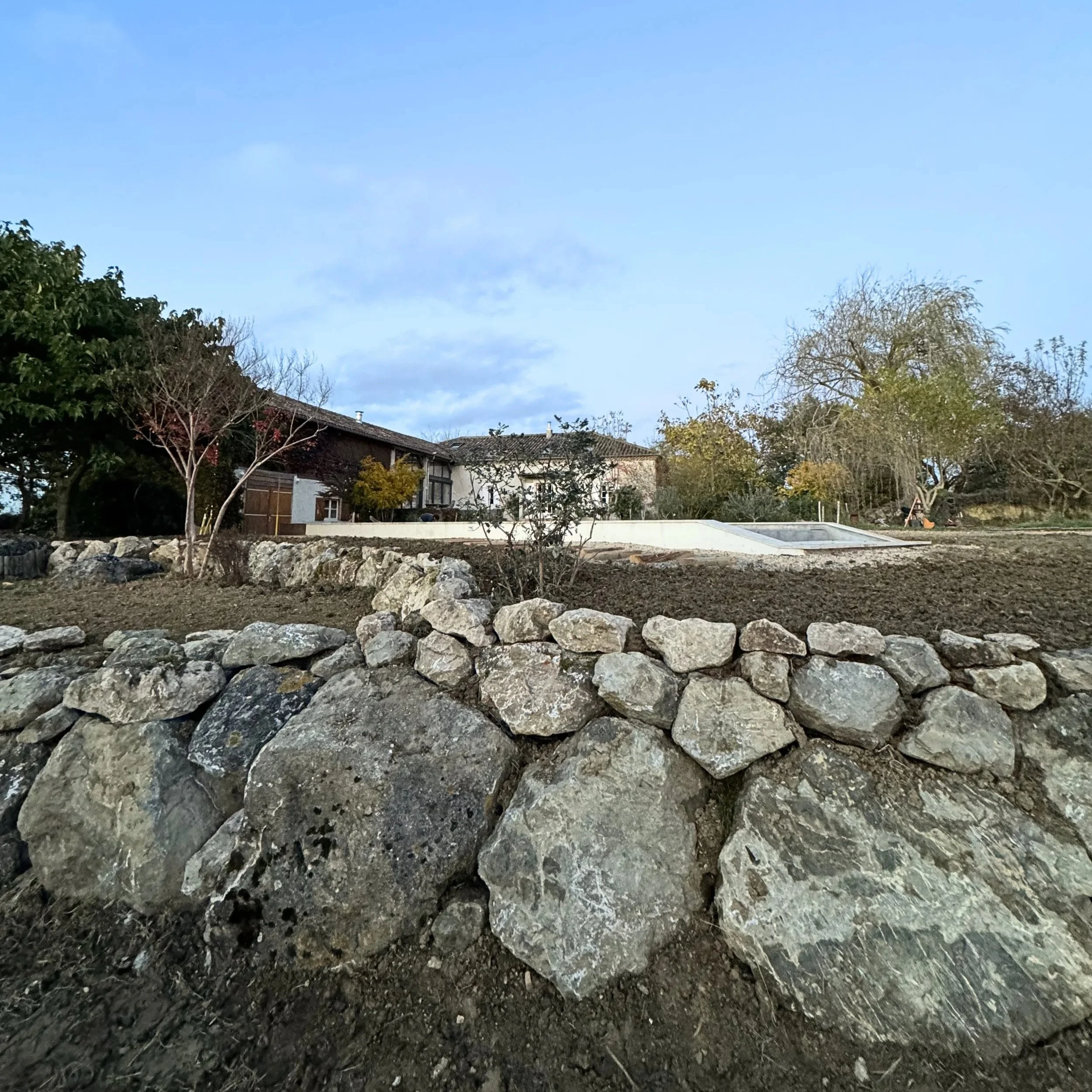 Un jardin avec une roche, de la terre nue, des arbres sans feuilles, une maison en arrière-plan, ciel bleu avec quelques nuages