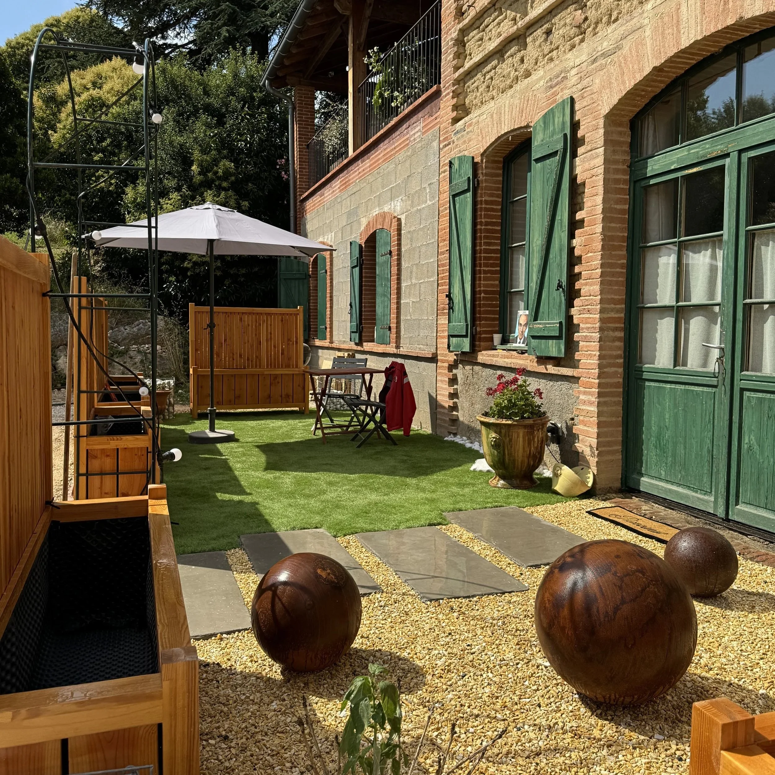 Jardin extérieur avec maison en pierre, fenêtres vertes, parasol, plantes, terrain en gazon artificiel, balles en bois, clôture en bois.