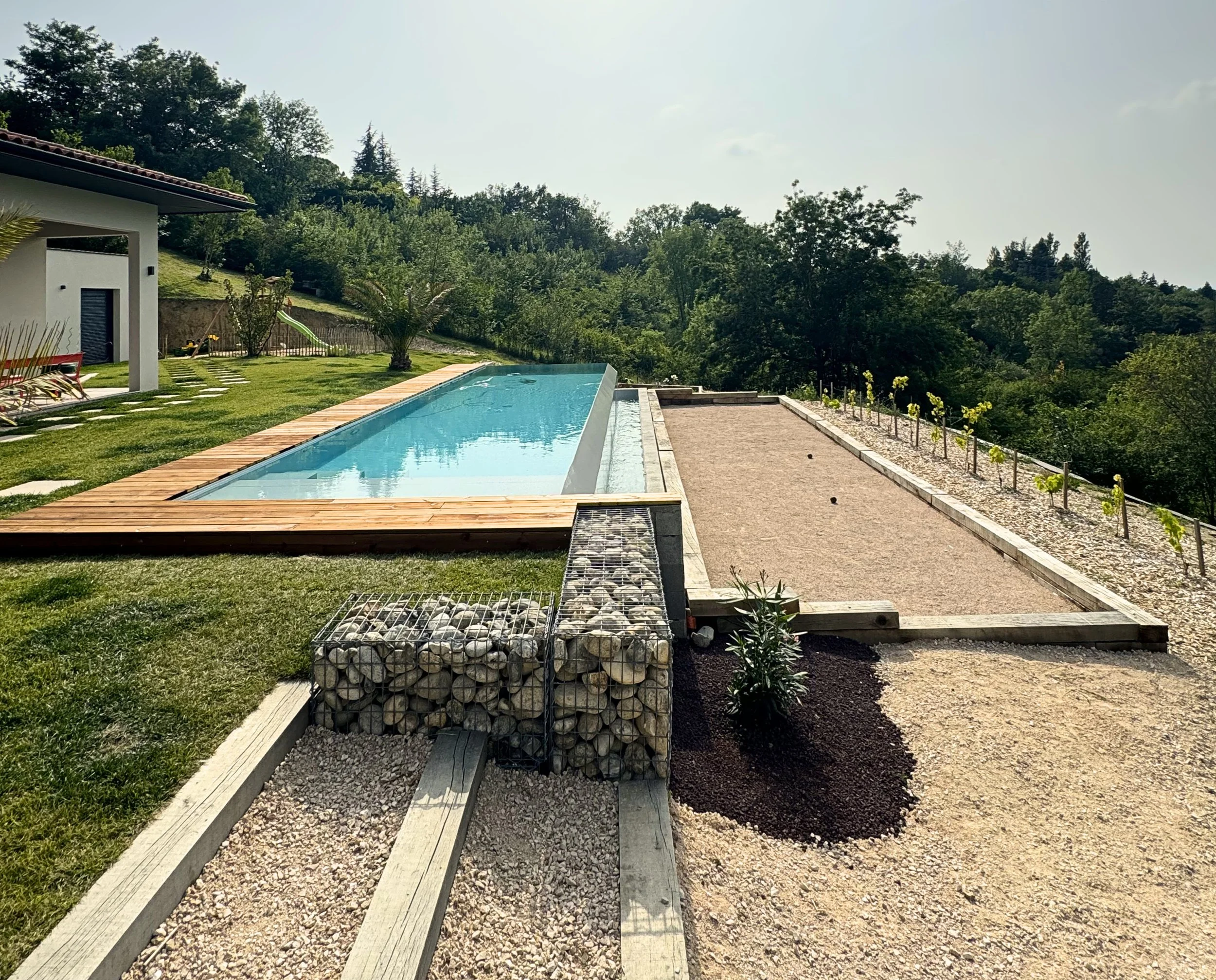 Une piscine à débordement dans un jardin entouré d'arbres, avec une maison moderne à gauche et une allée en gravier sur la droite.