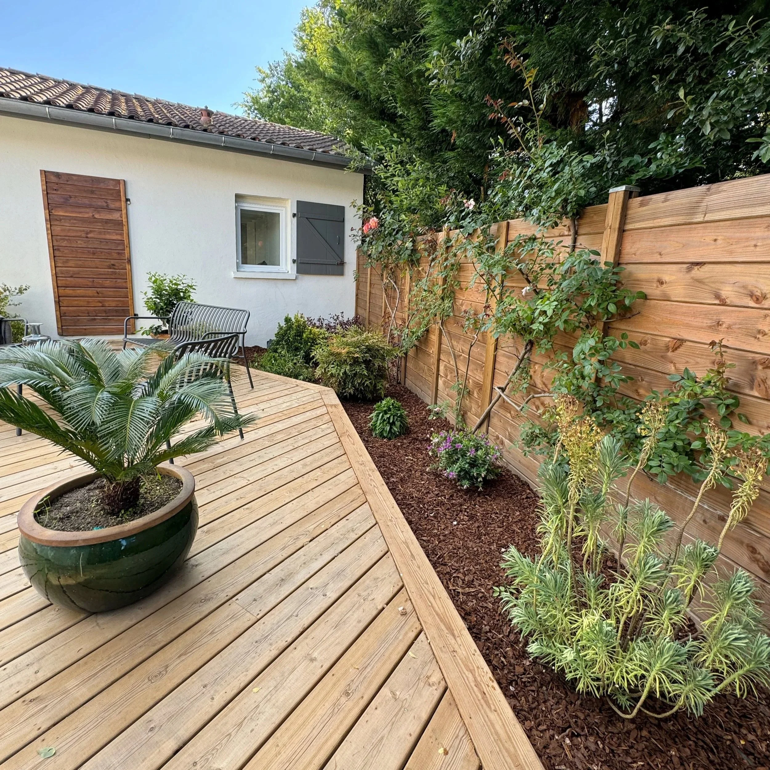 Jardin avec terrasse en bois, plantes vertes, clôture en bois, maison avec fenêtres et volets en bois, toiture en tuiles, ciel en arrière-plan.