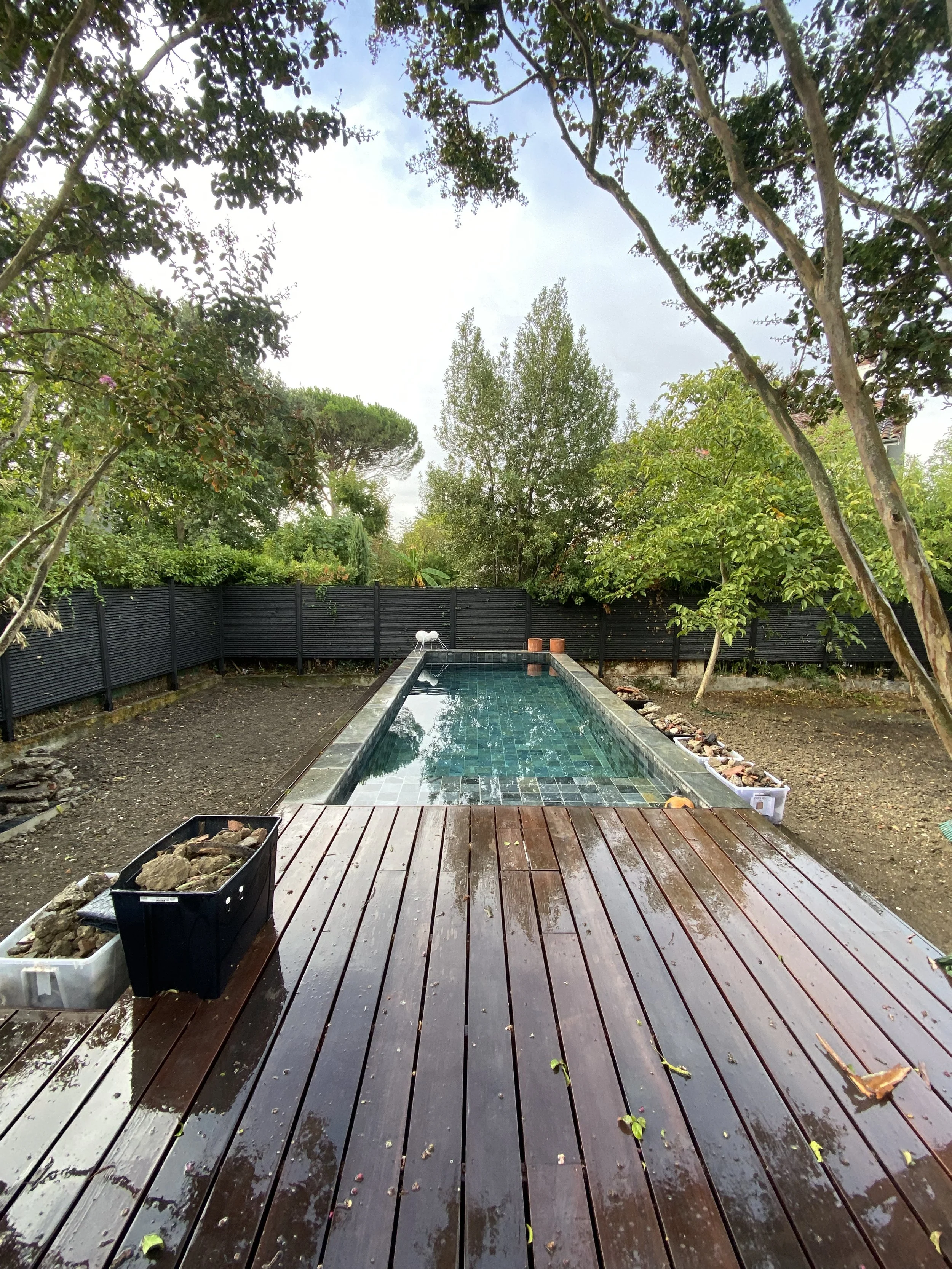 Un bassin de piscine rectangulaire avec un revêtement en mosaïque, entouré d'une terrasse en bois sombre mouillée, avec un fond d'arbres et un ciel nuageux.