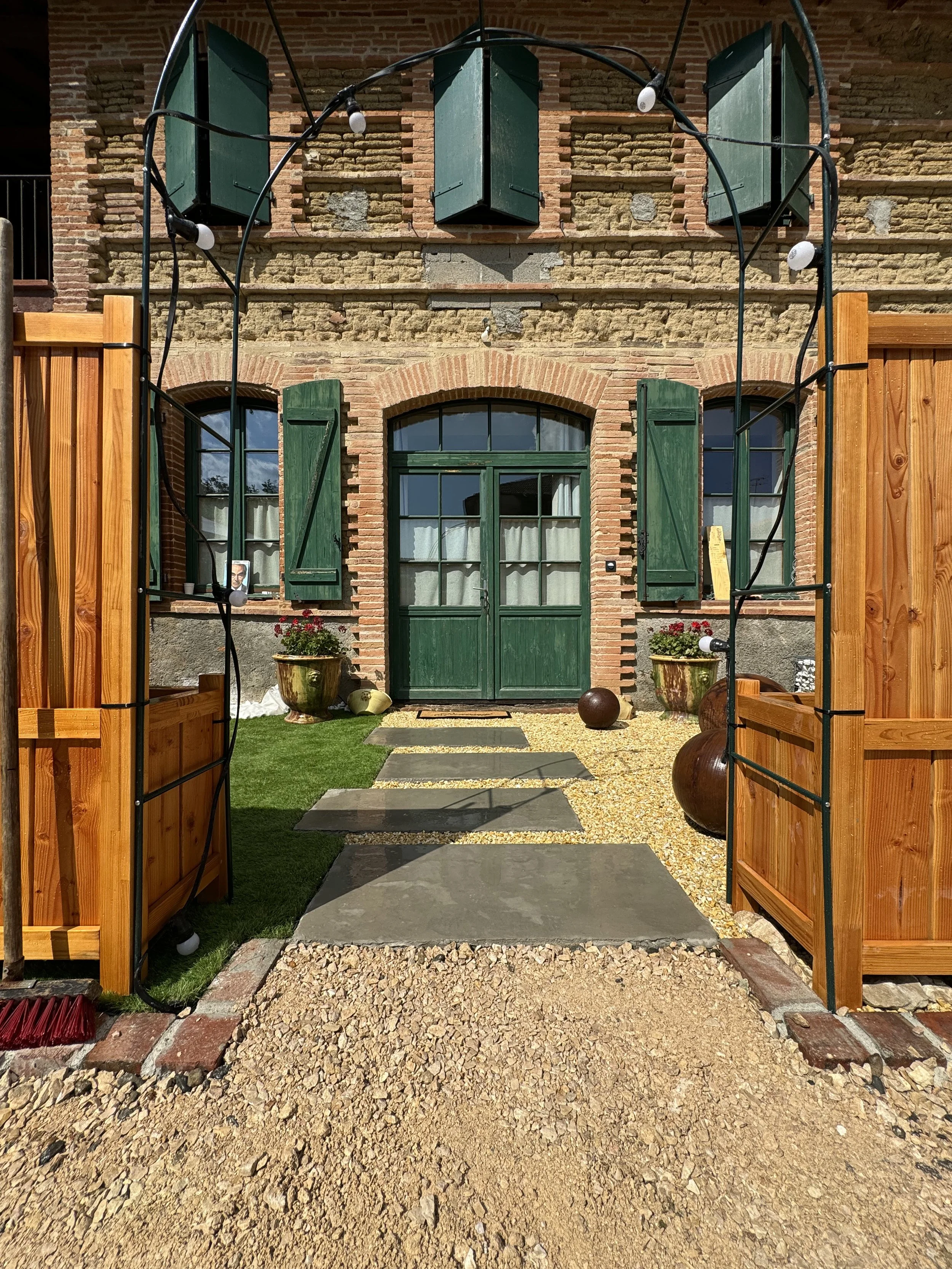 Vue d'une façade en briques avec fenêtres à volets verts, porte en acier vert, un escalier en pierre menant à la porte, entourée d'un jardin avec pelouse, plantes en pot et un chemin en gravier, encadré par une structure en bois et lumières extérieures.