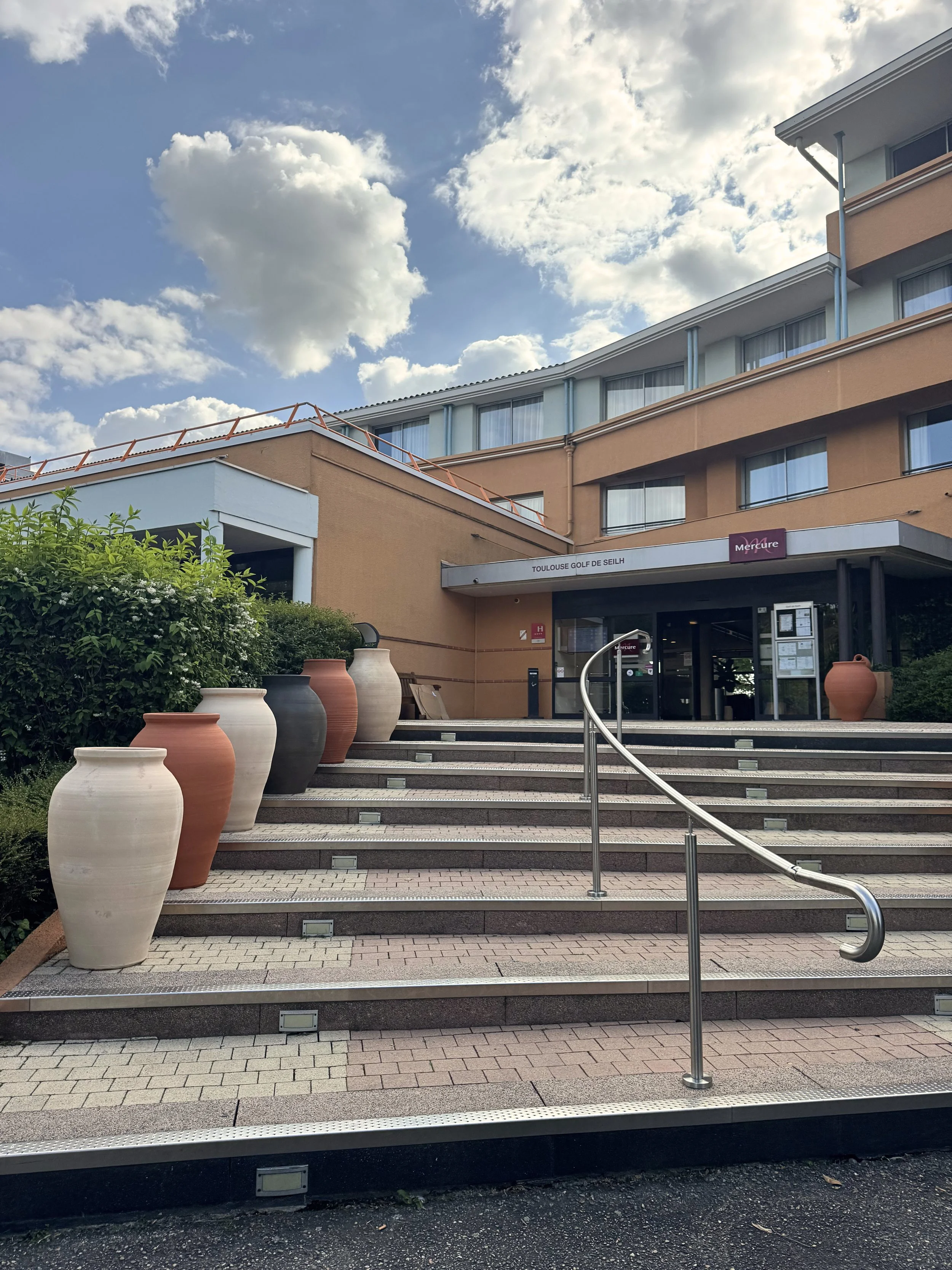 Façade d'un hôtel nommé 'Toulouse Golf De Seilh' avec un escalier en pierre, des pots en terre cuite, et un ciel nuageux.