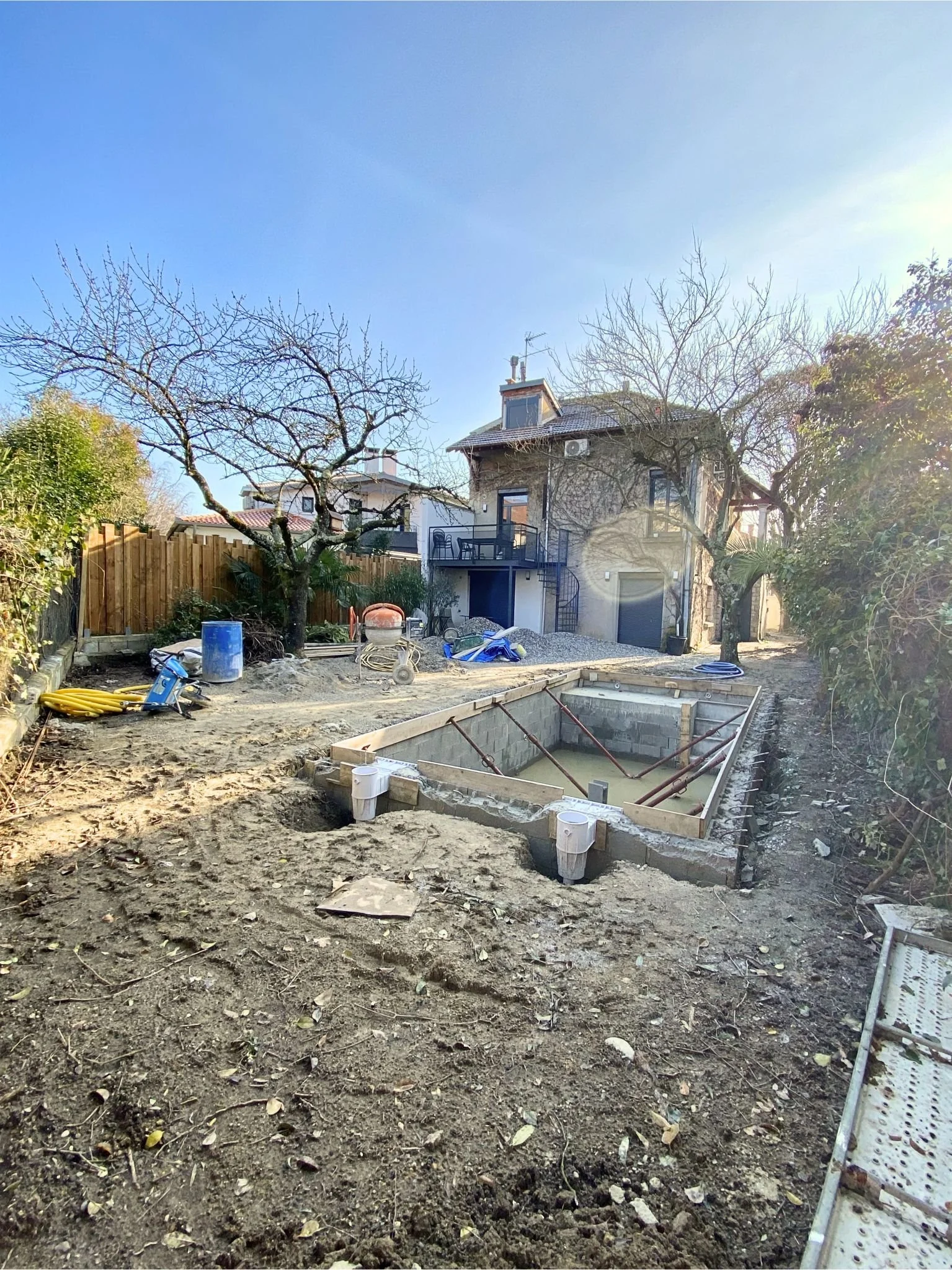 Chantier de construction dans un jardin arrière avec une piscine en creux, des outils, des tubes et des matériaux de construction autour. Arbres sans feuilles et maison en arrière-plan.