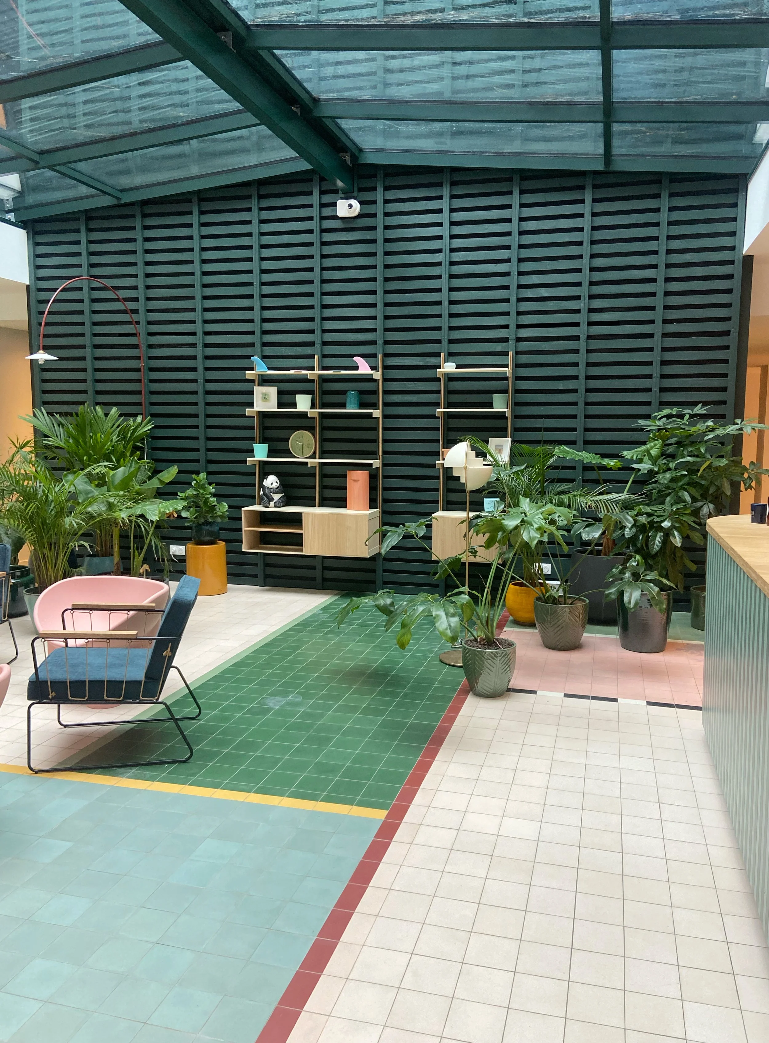 Intérieur d'une salle aux murs en bardeaux noirs, avec étagères en bois contenant des objets décoratifs, plantes vertes en pots, et un mobilier contemporain avec chaises, tapis et éclairages. Un plafond en verre permet à la lumière naturelle d'entrer.