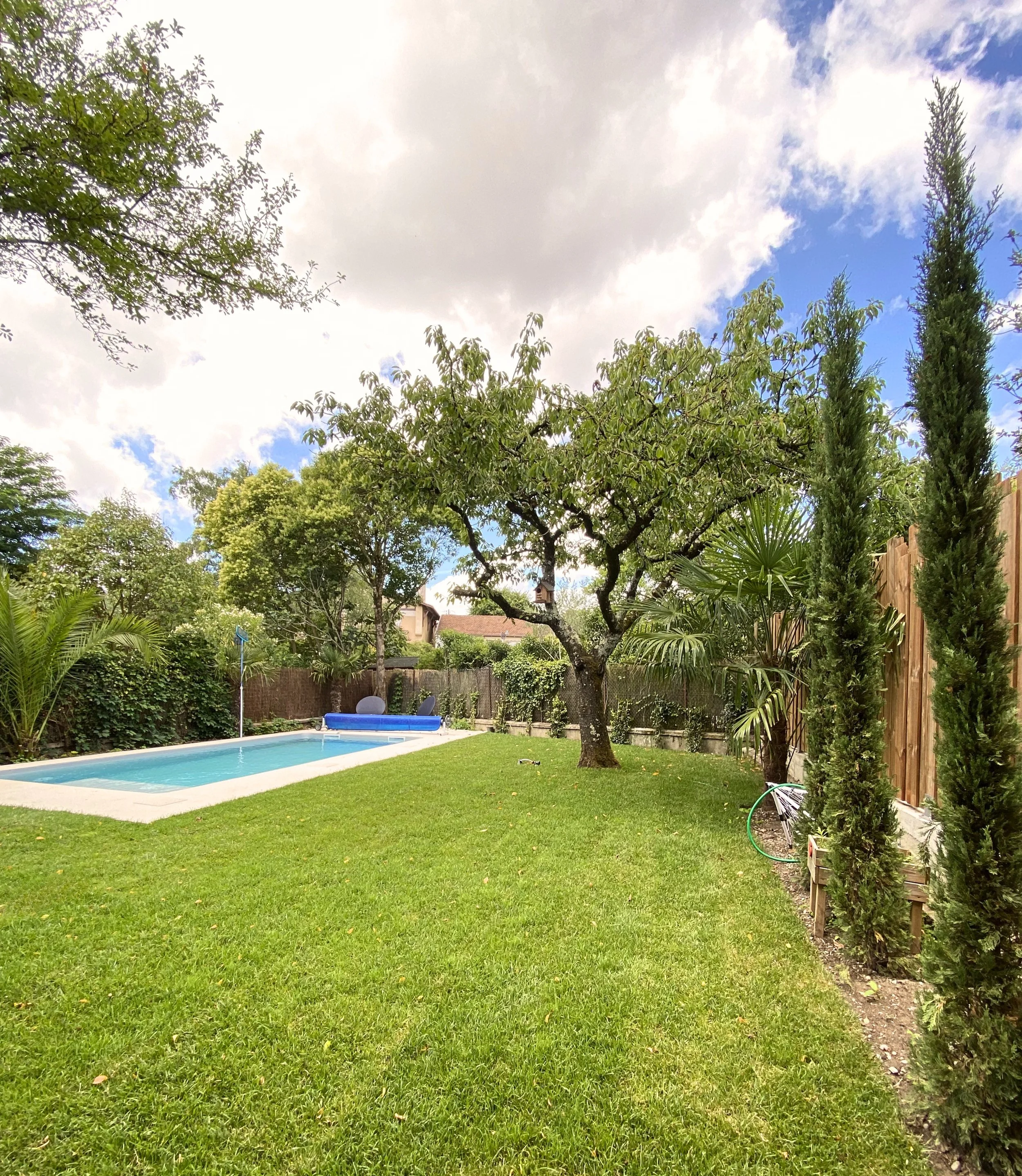 Jardin avec pelouse, arbres, piscine, clôture en bois, et ciel partiellement nuageux.