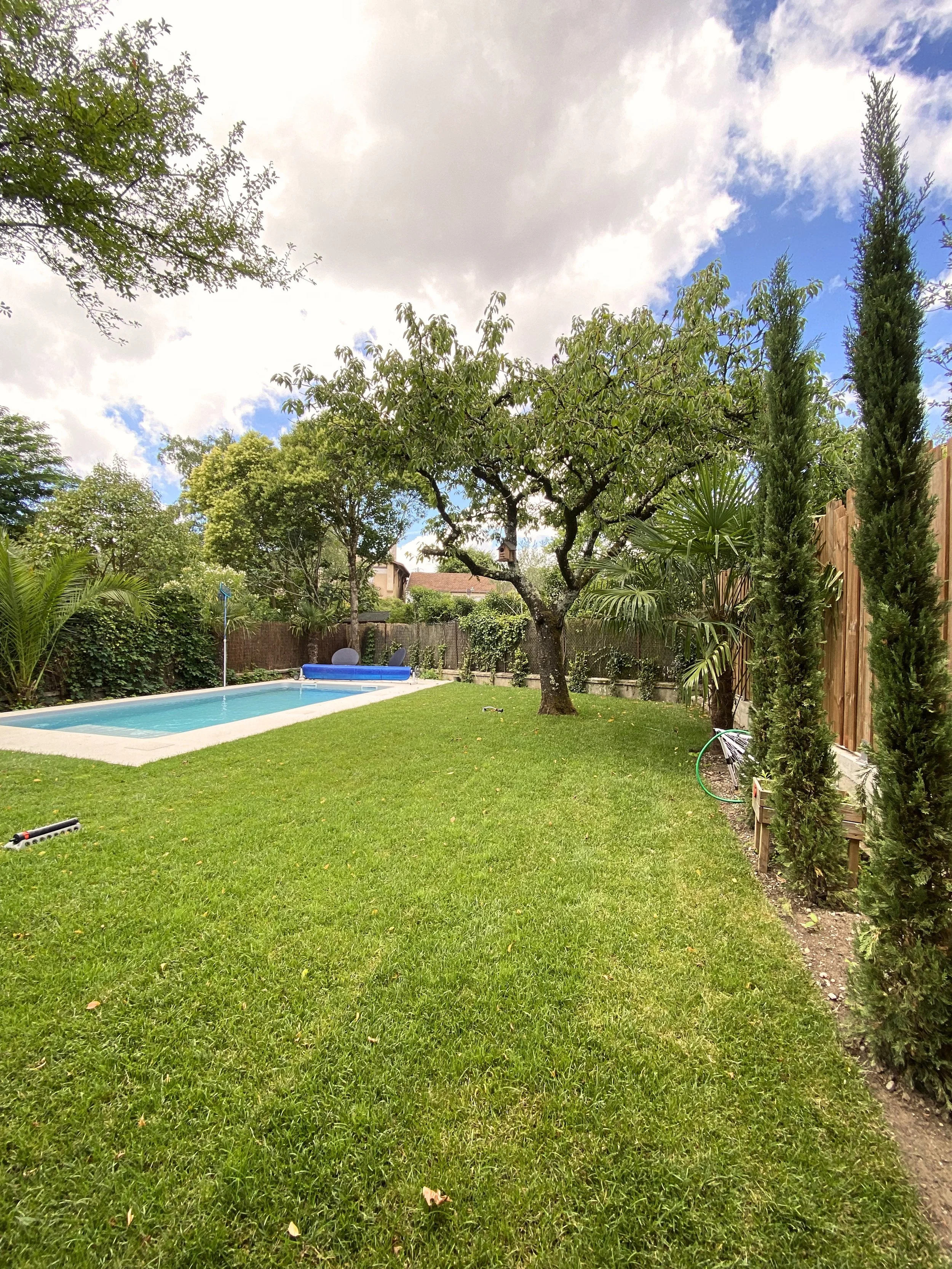 Une piscine dans un jardin entouré d'arbres et de haies, ciel partiellement nuageux.