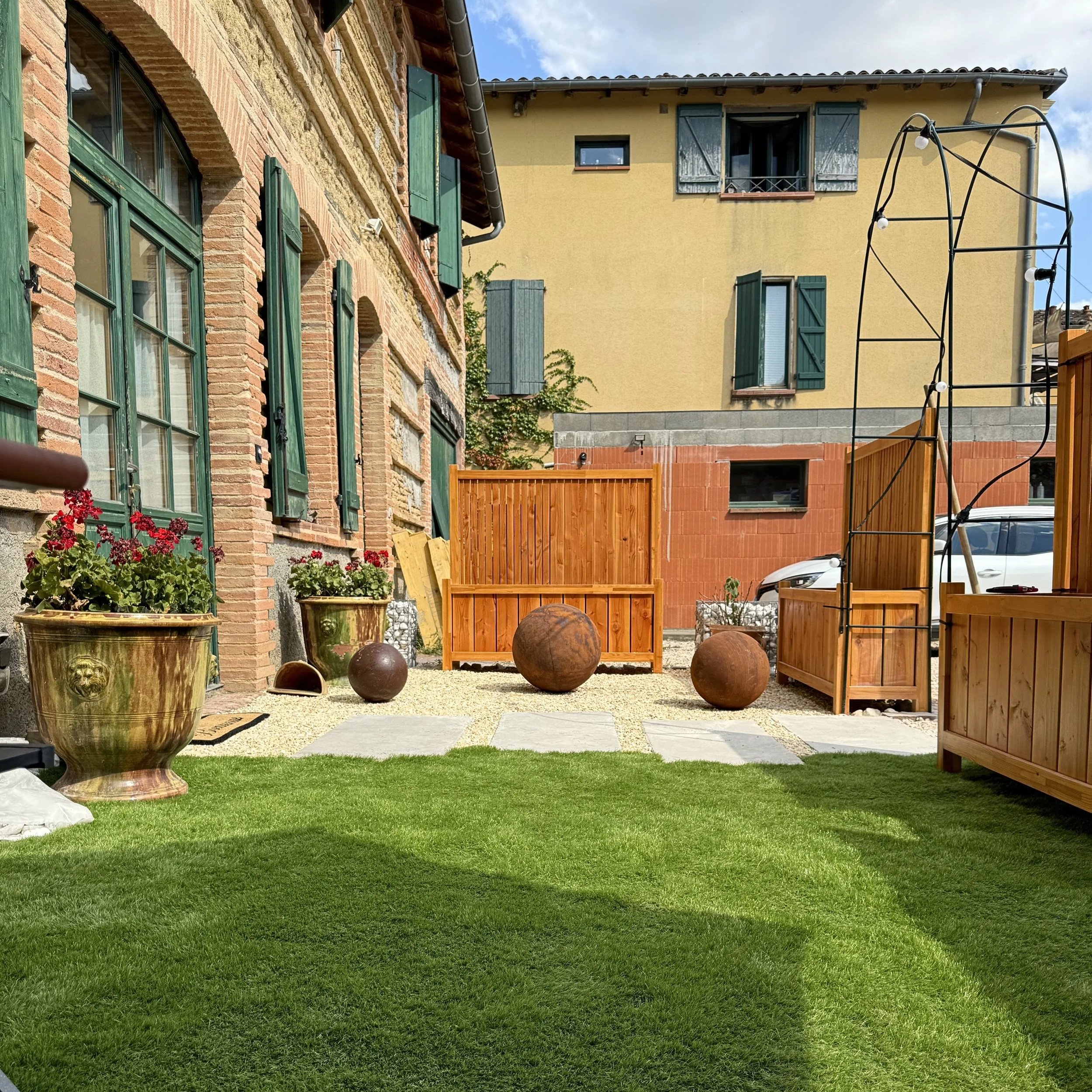 Jardin avec pelouse verte, sculptures en formes de boules en pierre, clôture en bois et maison en pierre avec volets verts sur fenêtres et portes.