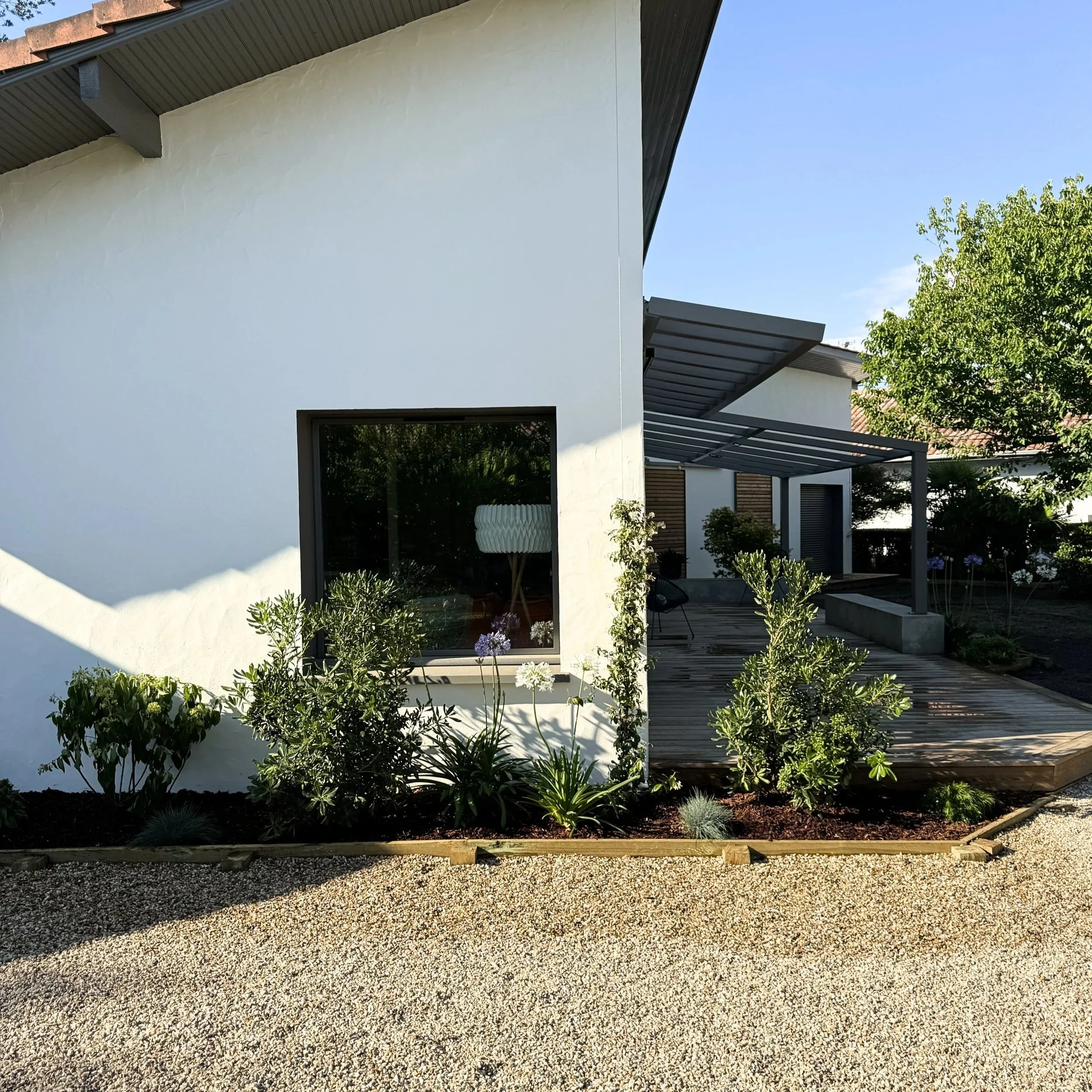 Une maison moderne avec une façade blanche, grandes fenêtres et une terrasse en bois, entourée de végétation et d'un jardin, sous un ciel clair.