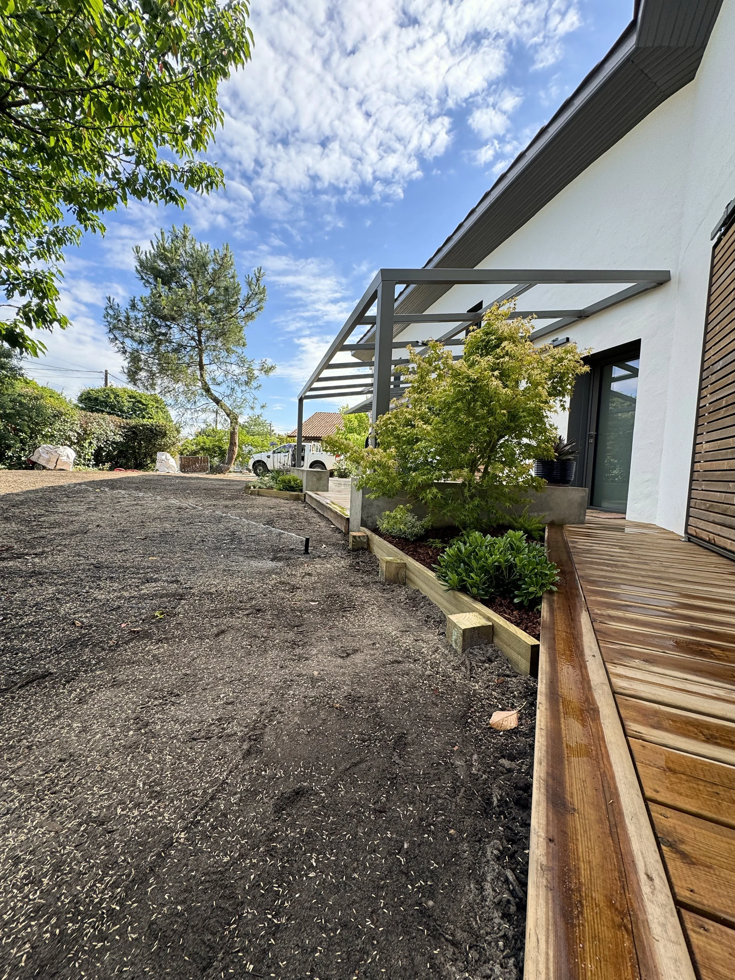 Vue d'une maison moderne avec une terrasse en bois, un arbre vert et un ciel partiellement nuageux.