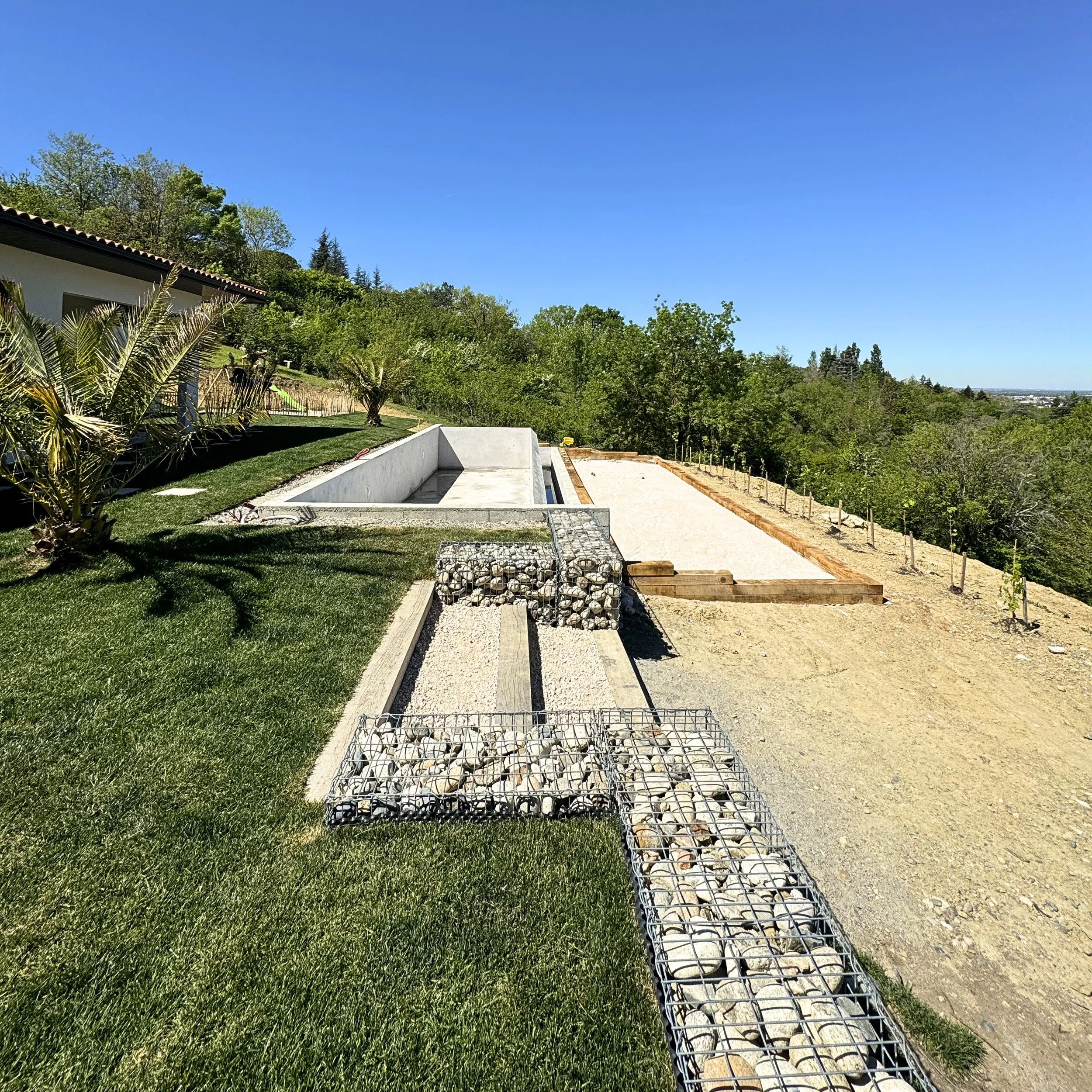 Un terrain en construction avec une piscine vide, des structures en béton, des fils métalliques, du gravier, des palettes en bois, entouré d'arbres et de verdure, sous un ciel bleu ensoleillé.