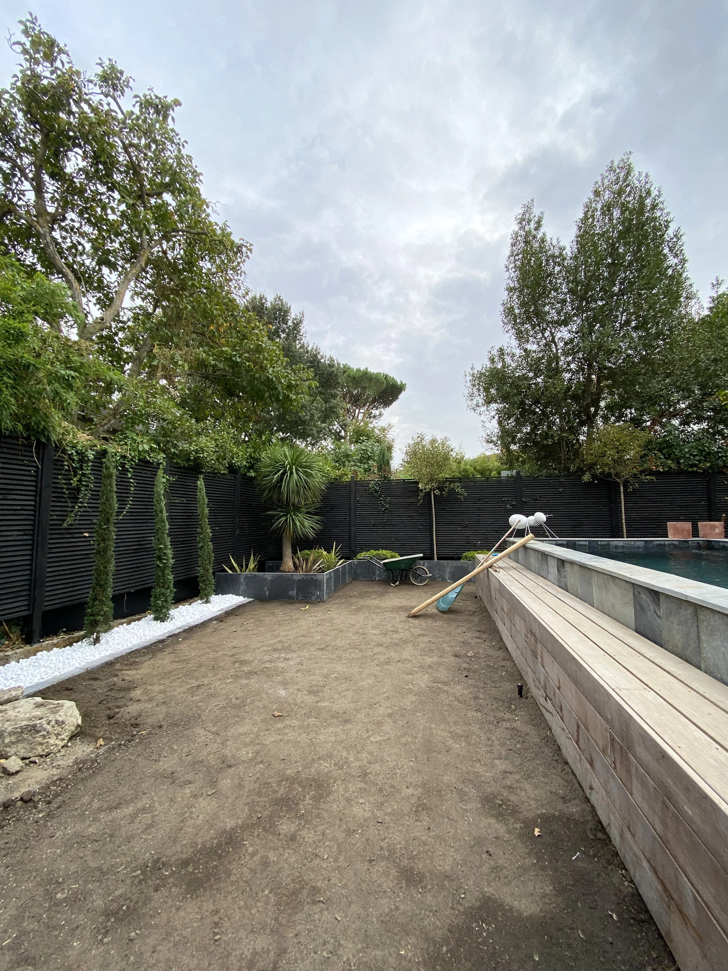 Jardinet avec des arbres, une clôture noire en arrière-plan, un bassin ou une piscine, et un sol en terre ou gravier. Il y a une pelle et un rouleau comme outils de jardinage. Le ciel est nuageux.