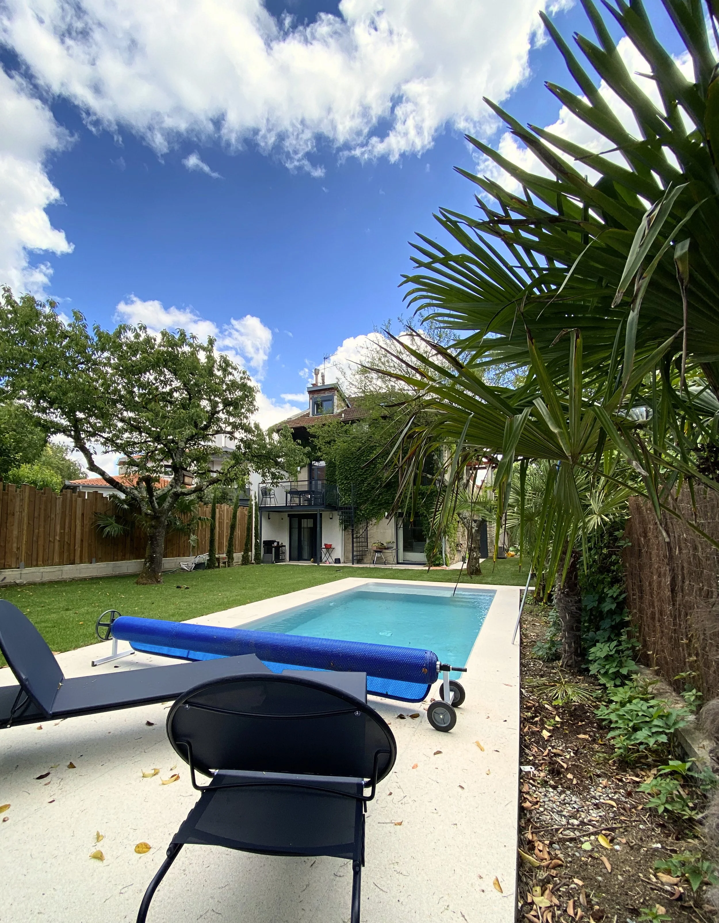 Piscine dans un jardin avec des chaises longues, entouré d'arbres et de plantes, sous un ciel bleu avec des nuages.