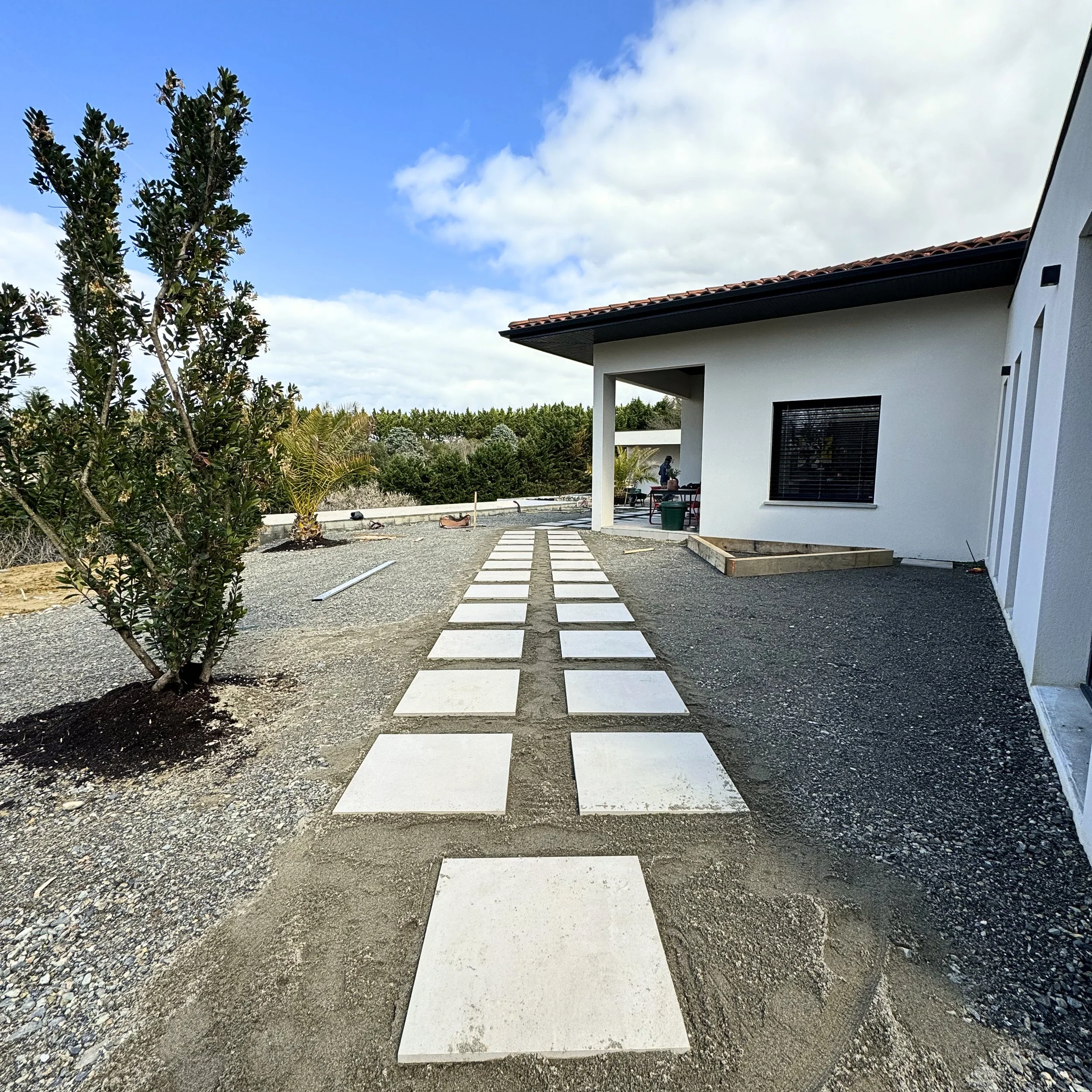 Travaux de construction avec des dalles en béton posées en ligne devant une maison moderne avec un toit en tuiles. Le ciel est bleu avec quelques nuages et il y a des arbres à l'arrière-plan.