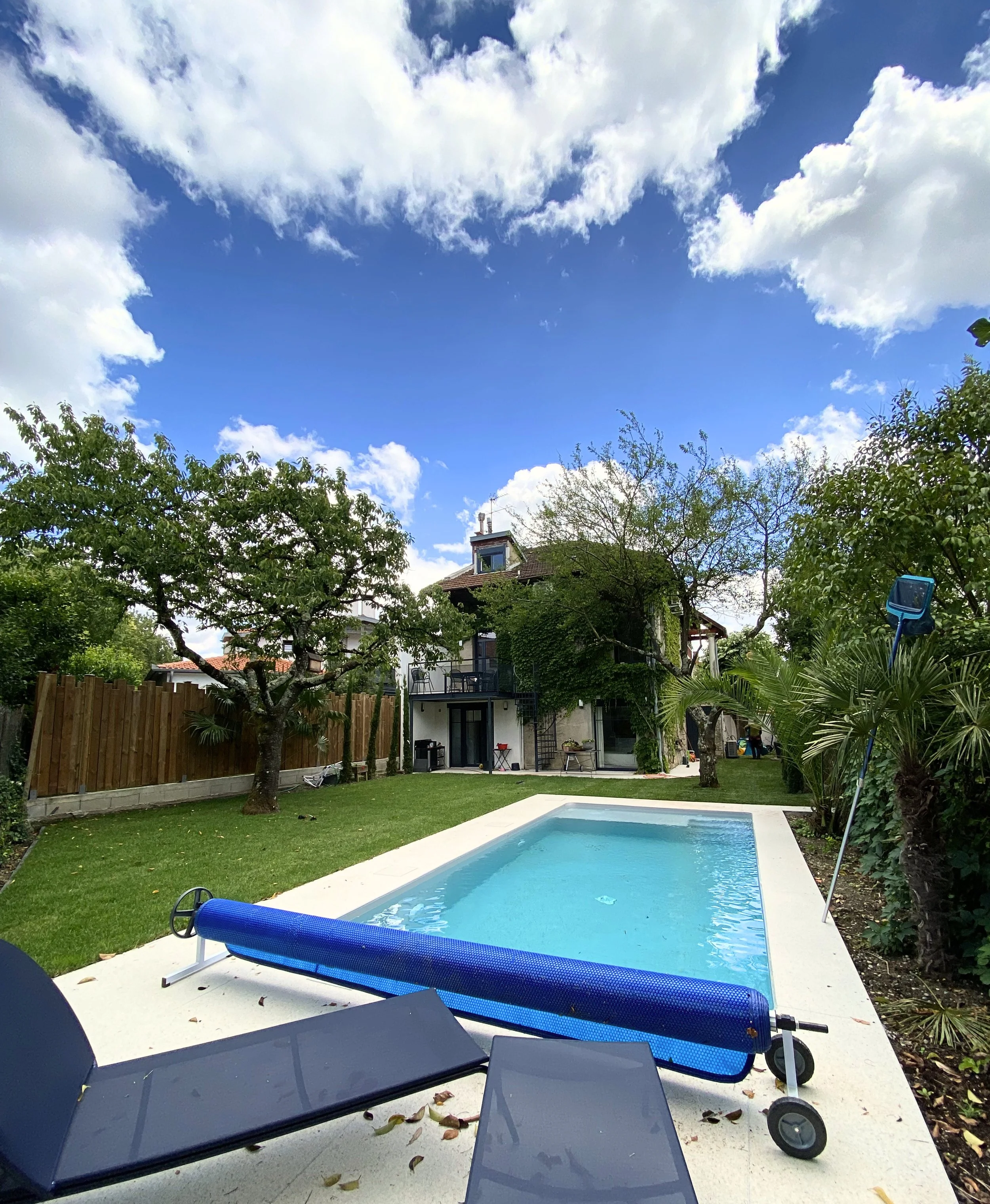 Une piscine dans un jardin entouré d'arbres, avec un meuble de jardin et une piscine à roulette en première plan, un bâtiment en arrière-plan, un ciel avec des nuages.