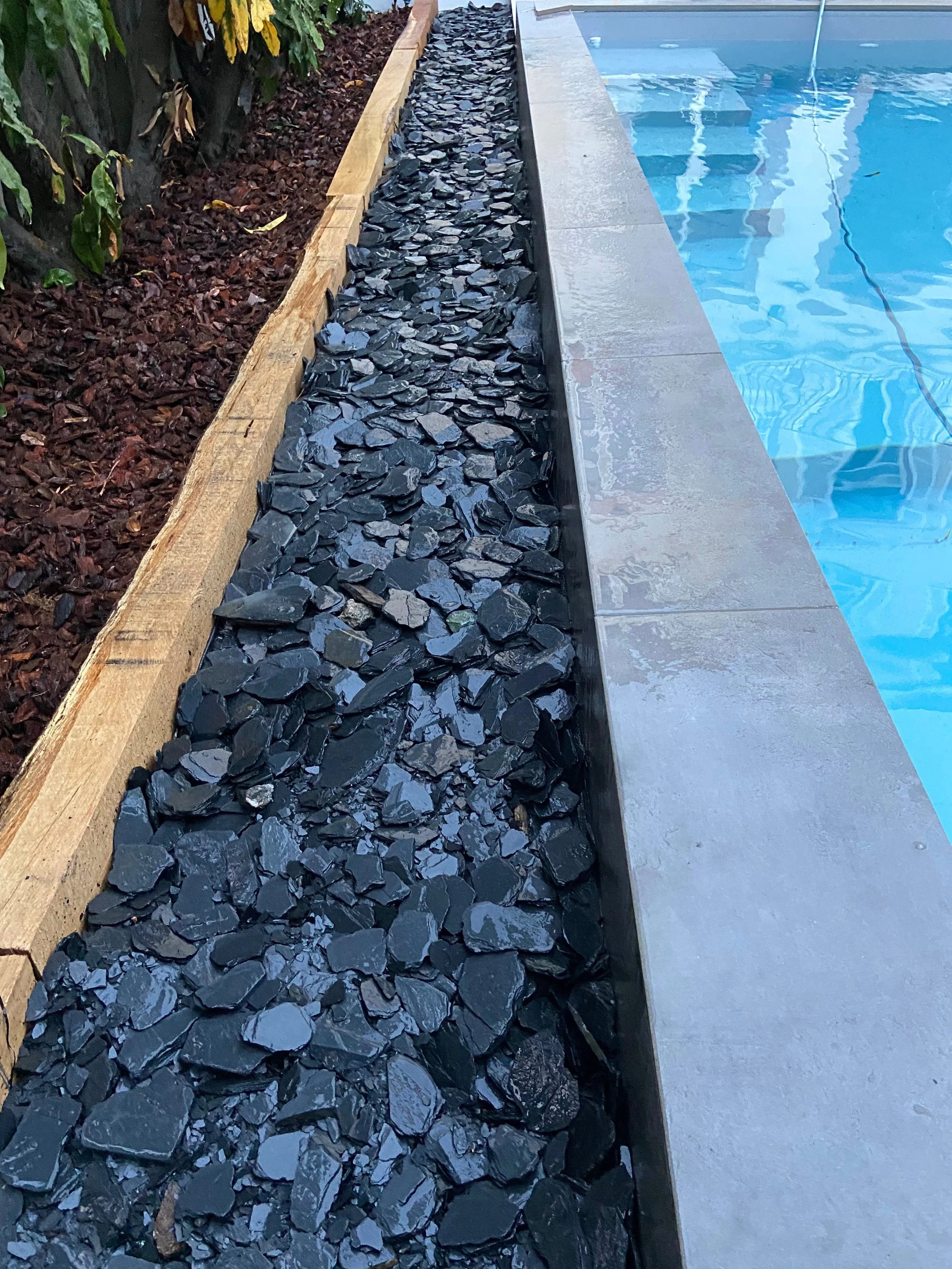 Gros plan d'une allée bordée de galets noirs, d'une terrasse et d'une piscine en arrière-plan.
