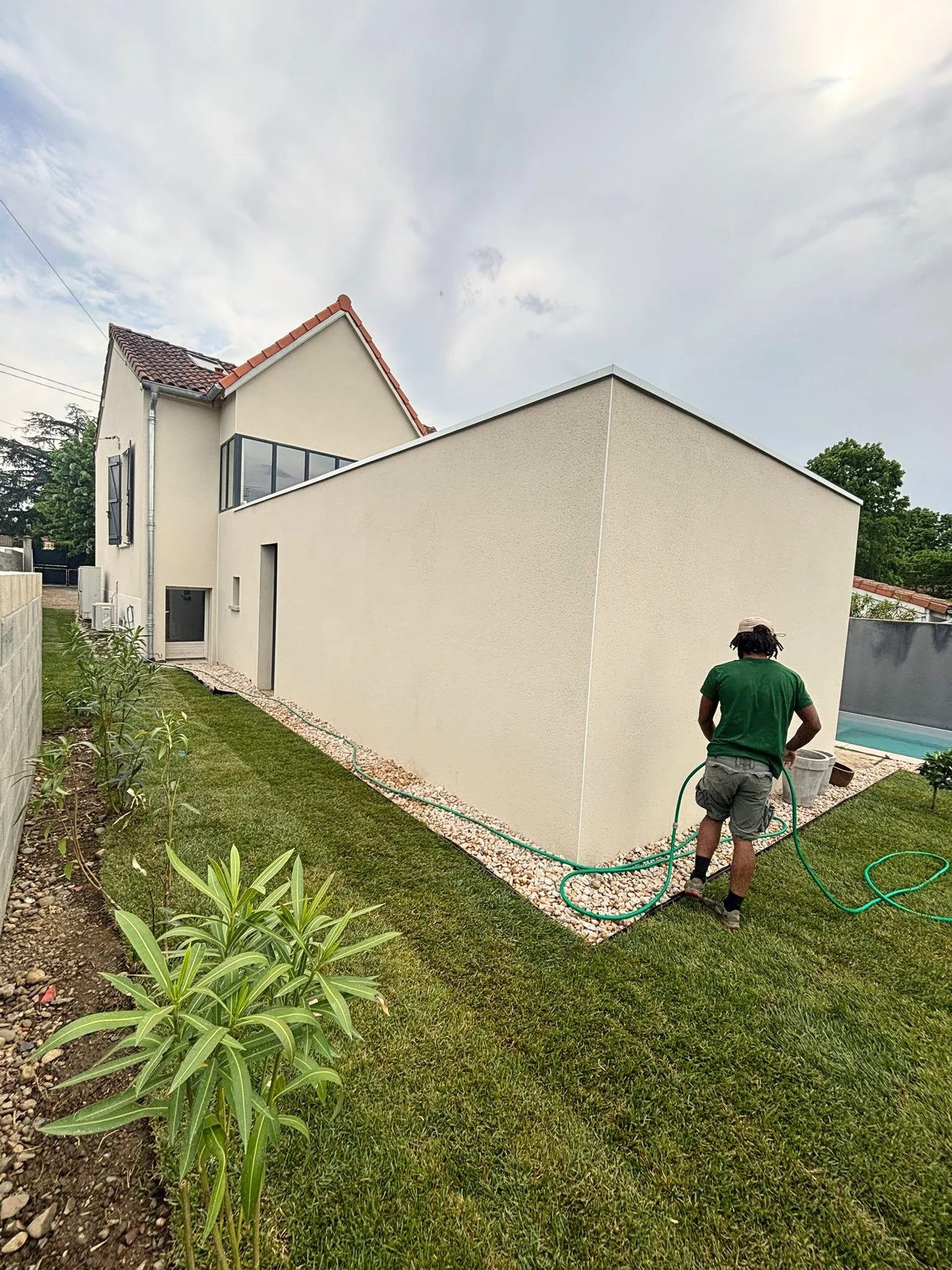 Un homme en chemise verte arrose un jardin avec un tuyau d'arrosage devant une maison moderne.