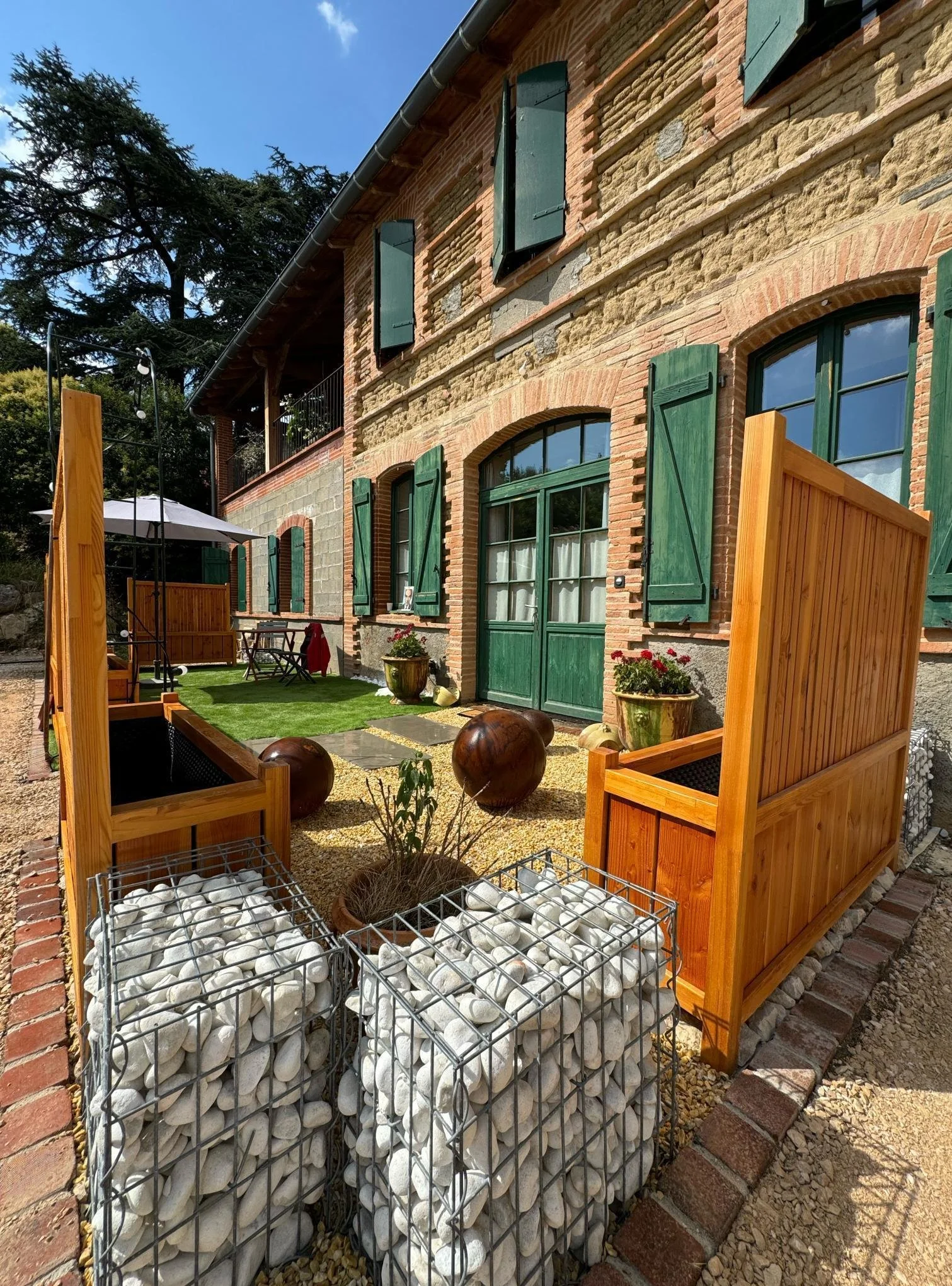 Jardin avec clôture en bois, plantes en pot, balcon à l'étage, maison en pierre avec volets verts, parasol, meubles extérieurs et terrain en pelouse.