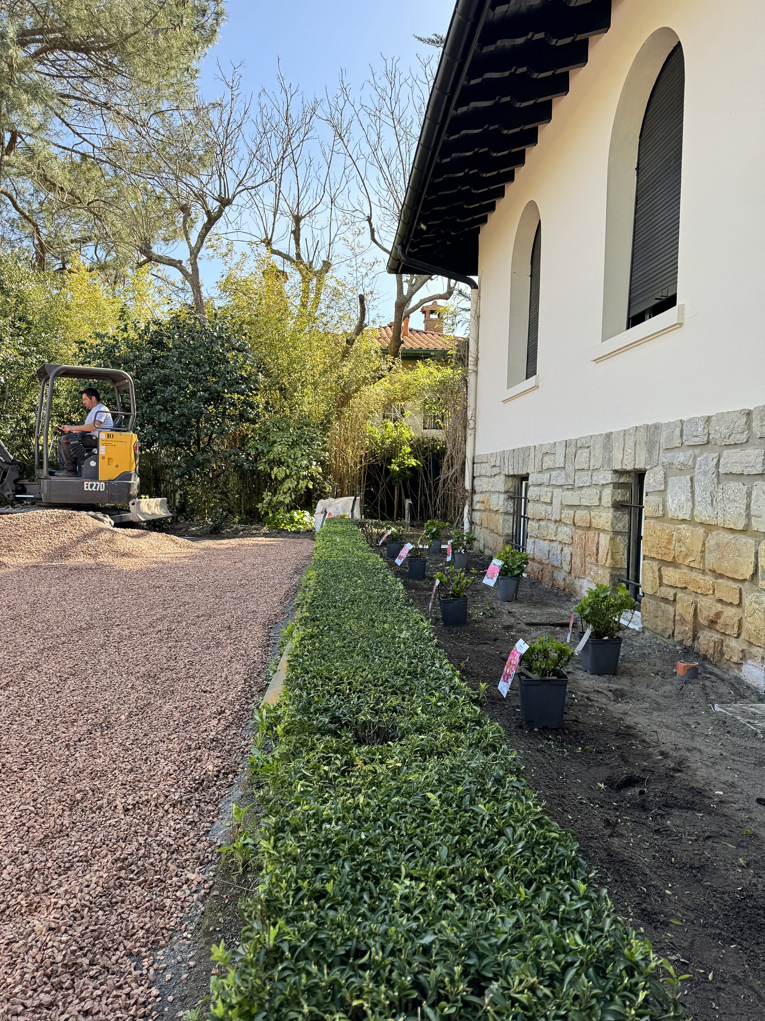 Une maison blanche avec des fenêtres à volets noirs, un chantier de jardinage avec des plantes en pot et un homme opérant une mini-pelle.
