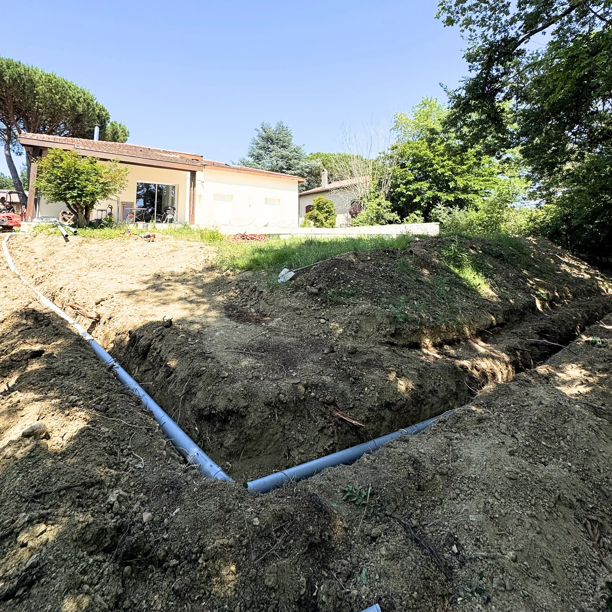 Une crevasse découverte dans le sol avec deux tuyaux en PVC bleu reposant dans la crevasse, entourée de terre et de végétation, avec une maison en arrière-plan et un ciel clair.