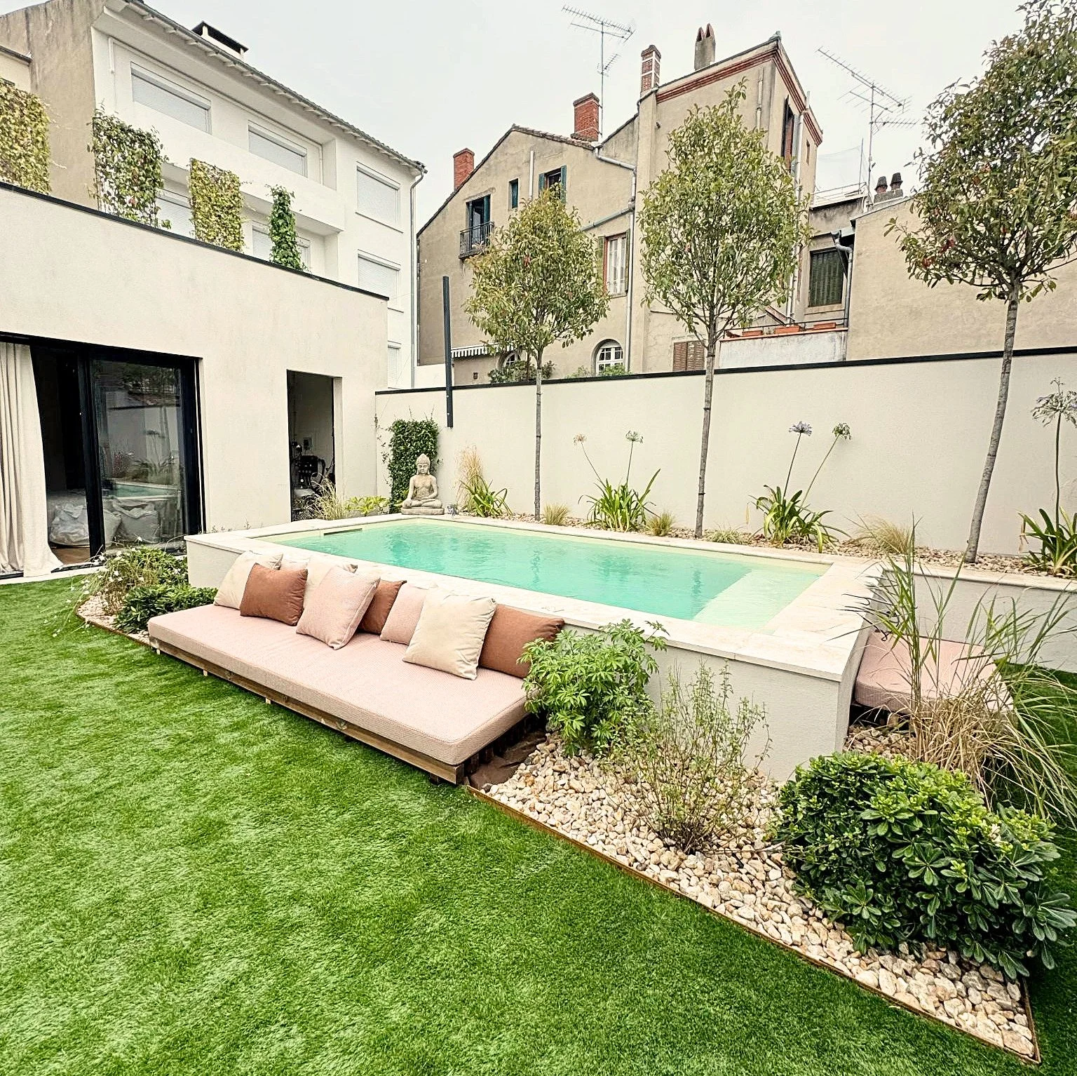 Cour arrière avec piscine, canapé avec coussins, plantes, arbres, mur blanc, maisons en arrière-plan.