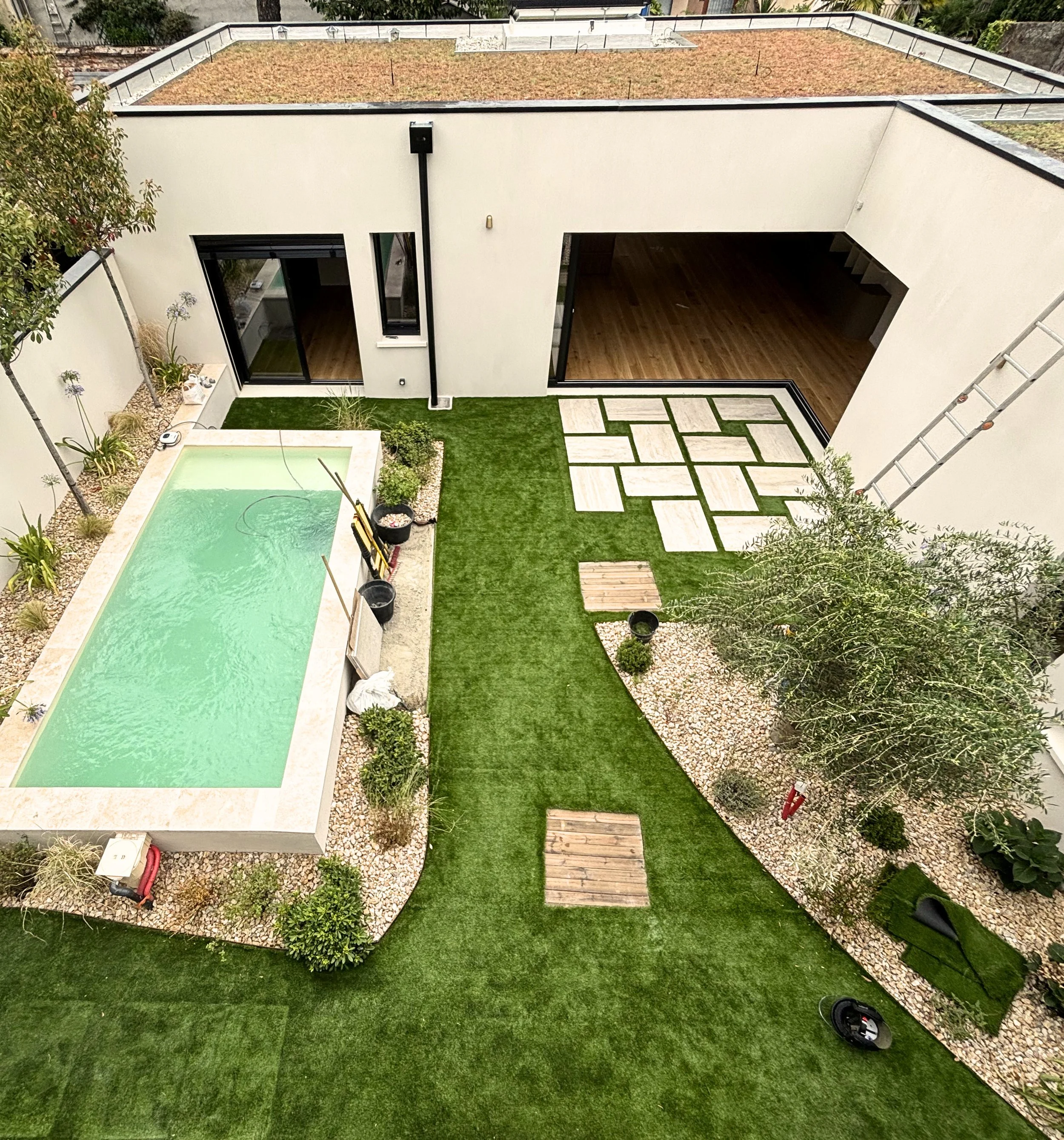 Vue aérienne d'une cour intérieure avec une piscine, des plantes, des chemins pavés, une maison moderne avec espace intérieur visible, et un ciel clair.
