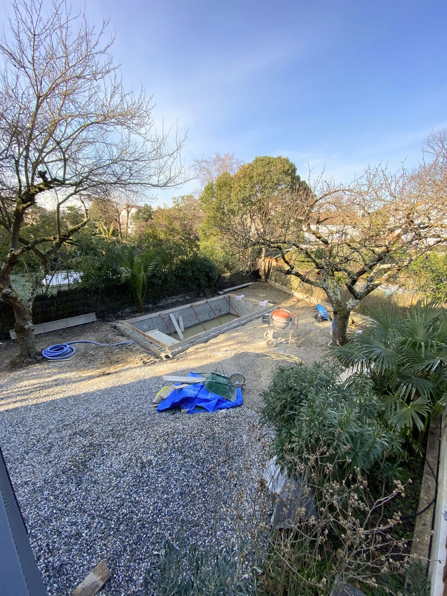 Jardin avec arbres, végétation, sol gravillonné, piscine en construction, matériel de chantier, ciel bleu.