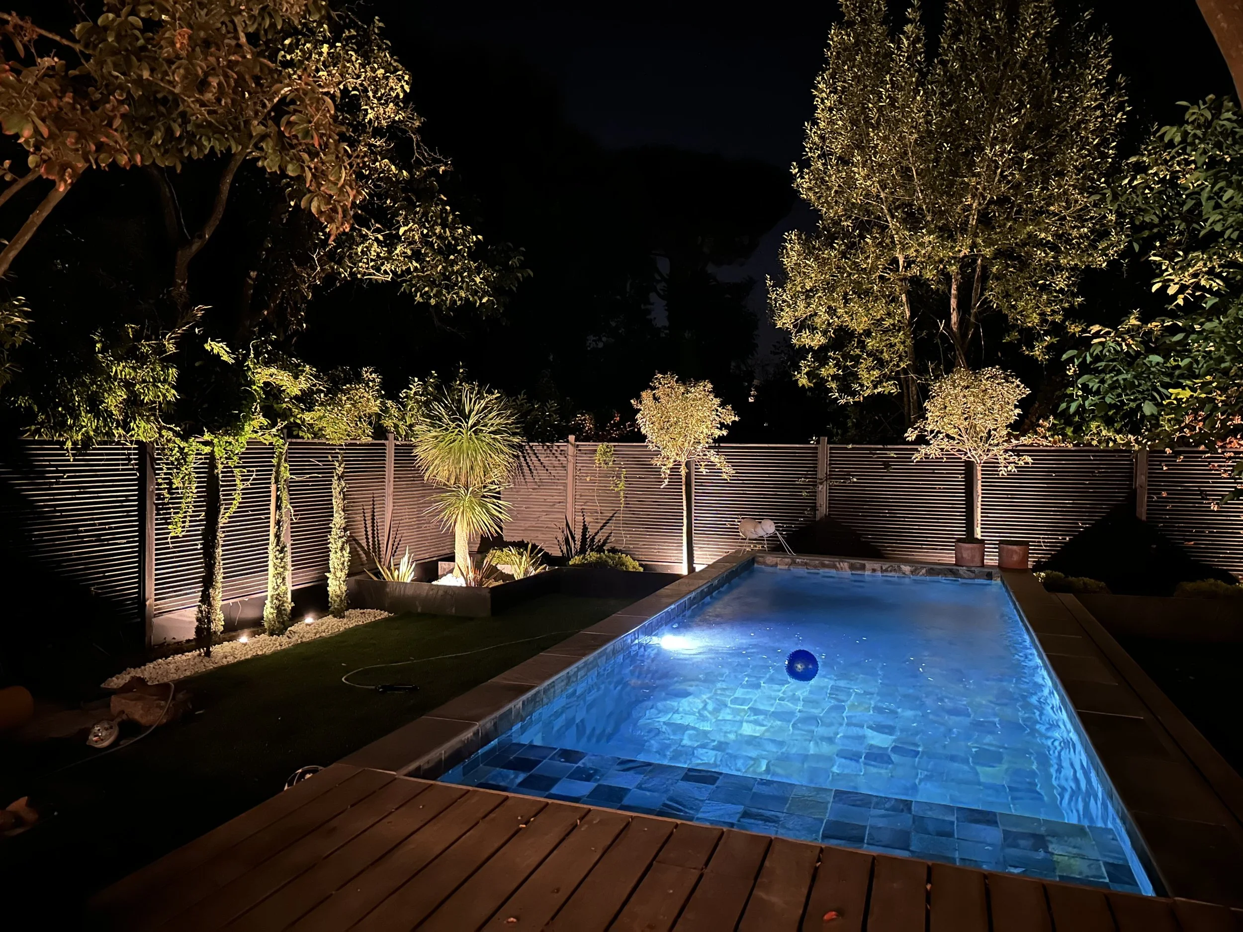 Une piscine éclairée la nuit, entourée de buissons et d'arbres, avec un miroir d'eau dans un jardin privé.