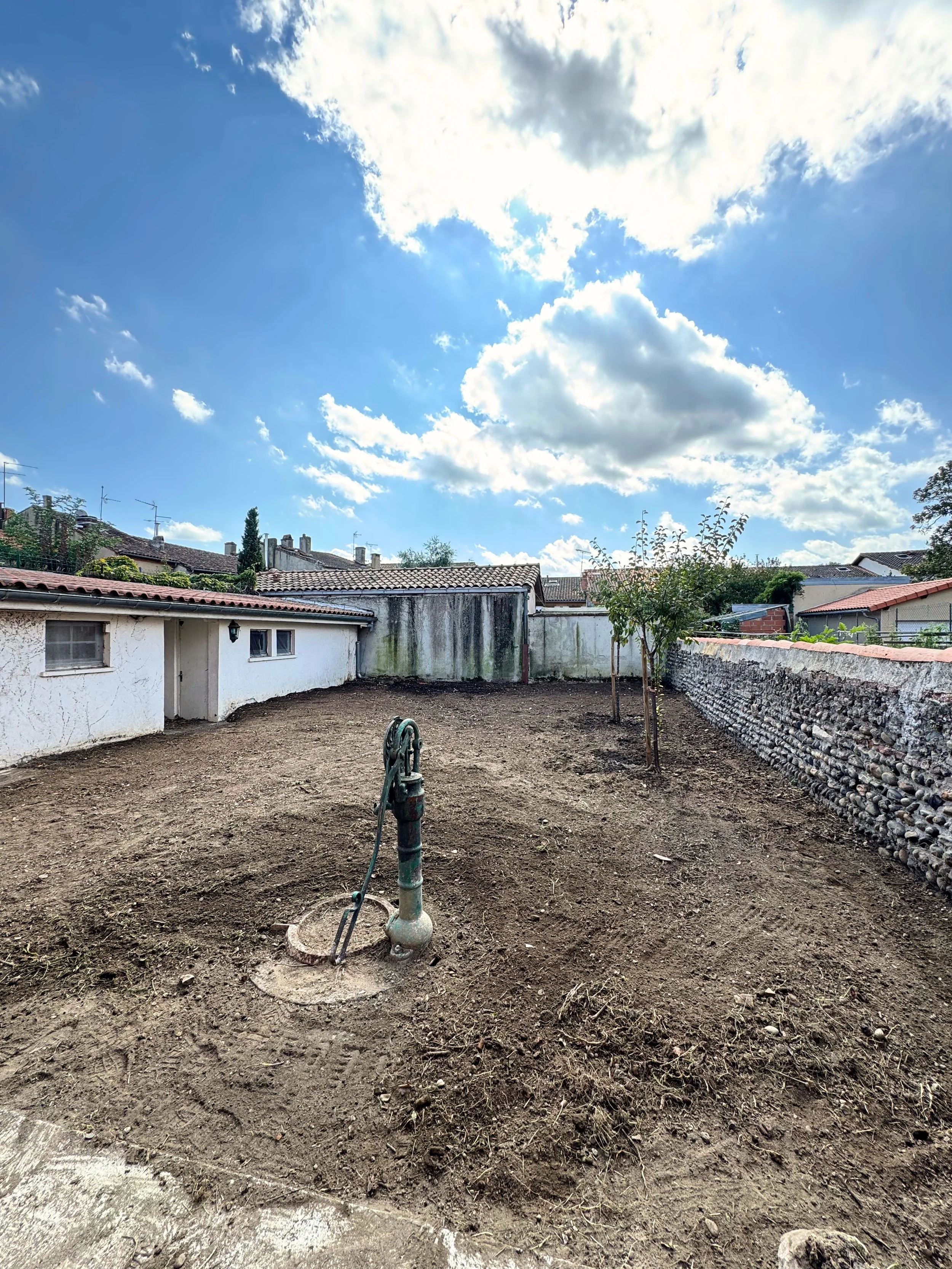 Un jardin en cours de construction avec le sol préparé, deux jeunes arbres plantés, une vieille canalisation en métal, un mur en pierre, et des bâtiments résidentiels avec des toits en tuiles sous un ciel partiellement nuageux.