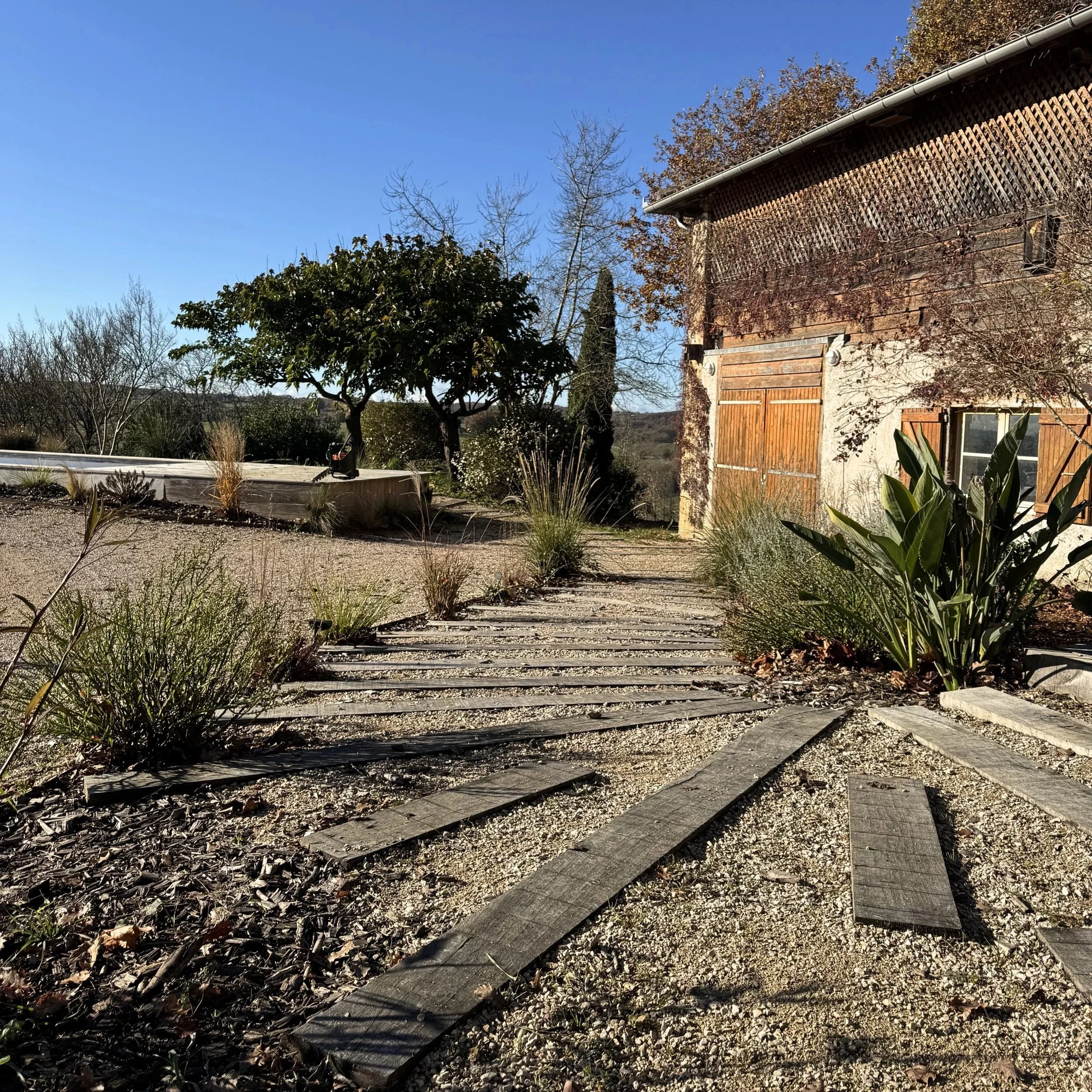 Chemin en bois dans un jardin, enceinte de plantes et d'arbres, bâtiment en pierre et bois, ciel bleu.