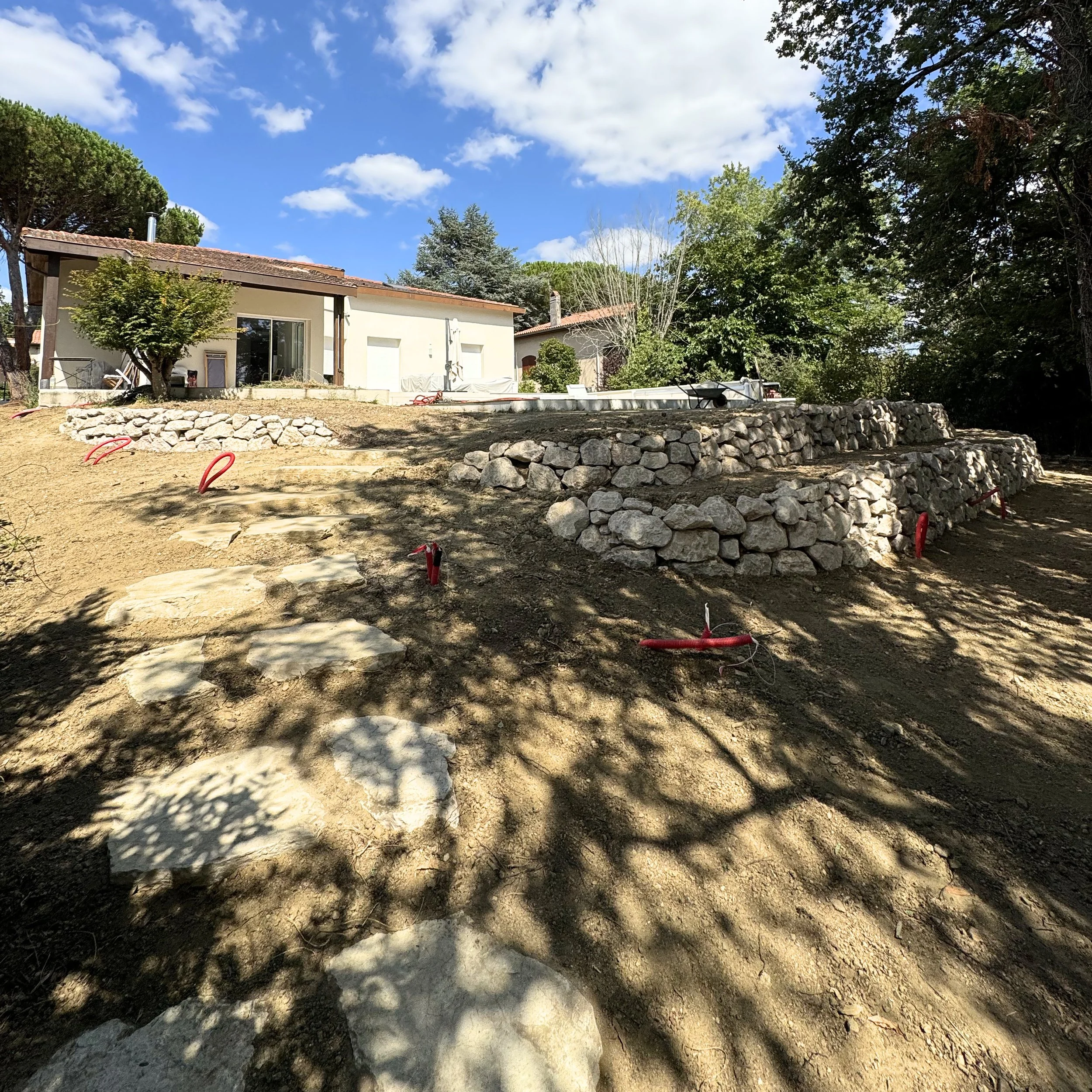 Une maison moderne entourée d'arbres avec un ciel bleu et quelques nuages, un terrain en cours d'aménagement avec des pierres et des tuyaux.