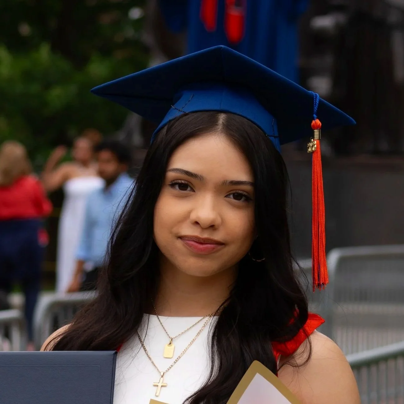 Young woman in graduation cap and gown smiling, with blurry people in the background.
