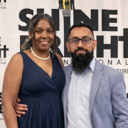 A smiling woman in a navy blue dress and a man in a light gray suit with glasses and a beard, standing together in front of a banner that reads 'SHINE A LIGHT' with additional text.
