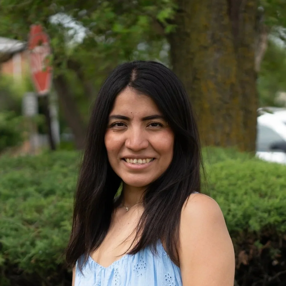 A woman with long black hair wearing a light blue sleeveless top, standing outdoors with trees and greenery in the background.