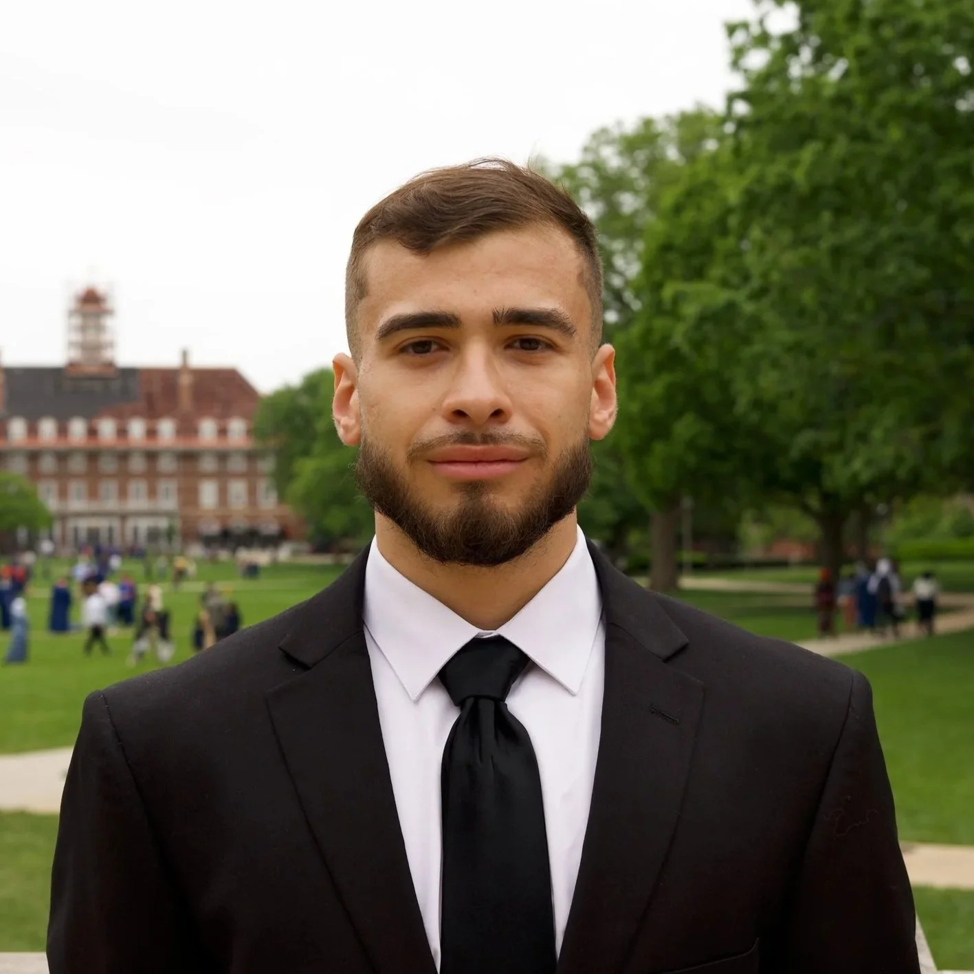 A young man with a beard and short brown hair, dressed in a black suit with a white shirt and black tie, standing outdoors in a park with green trees and a historic building in the background.