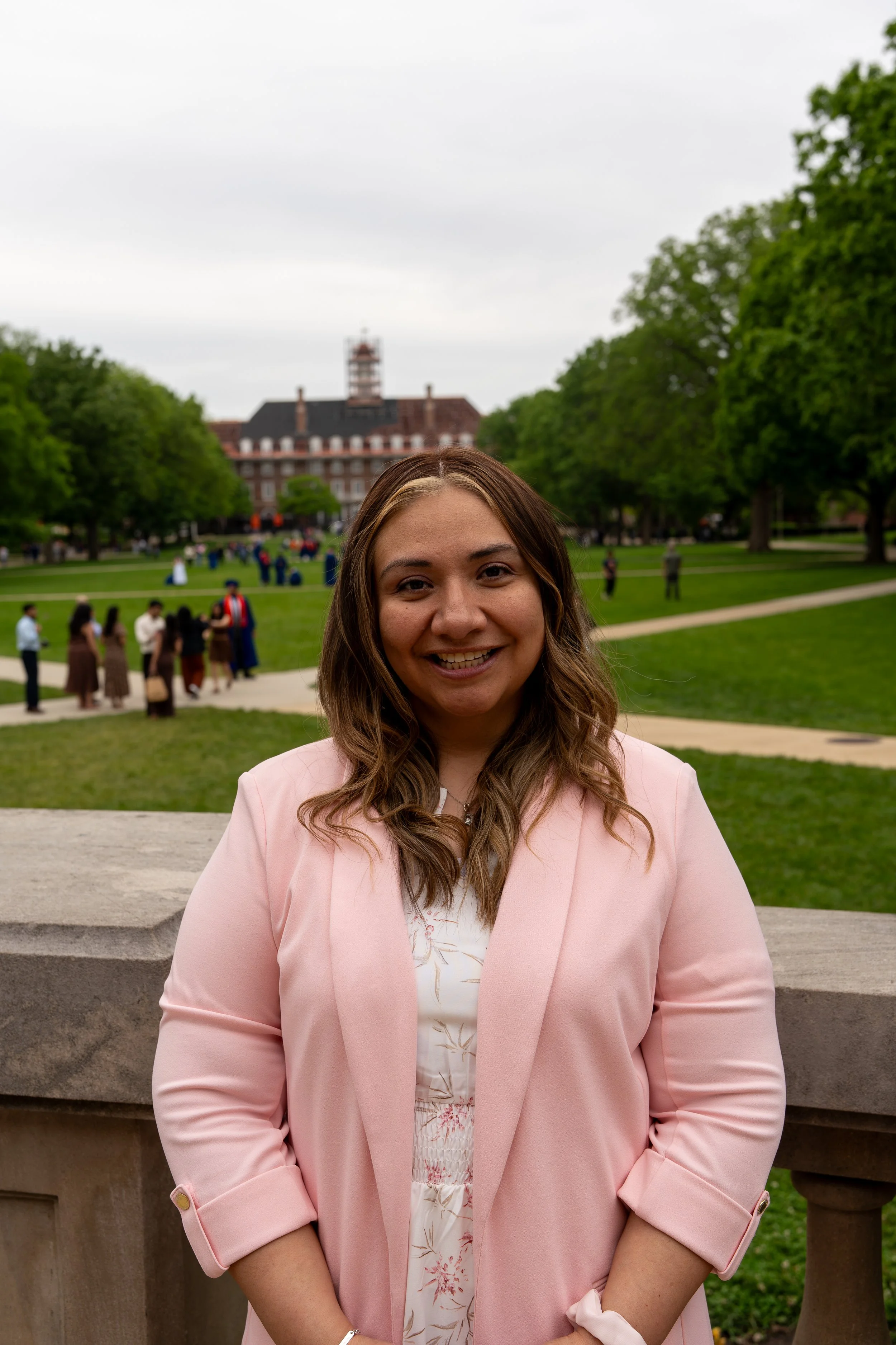 A woman with brown hair and a pink blazer smiling outdoors, with a park and a historic building in the background, and several people walking and gathering in the park.