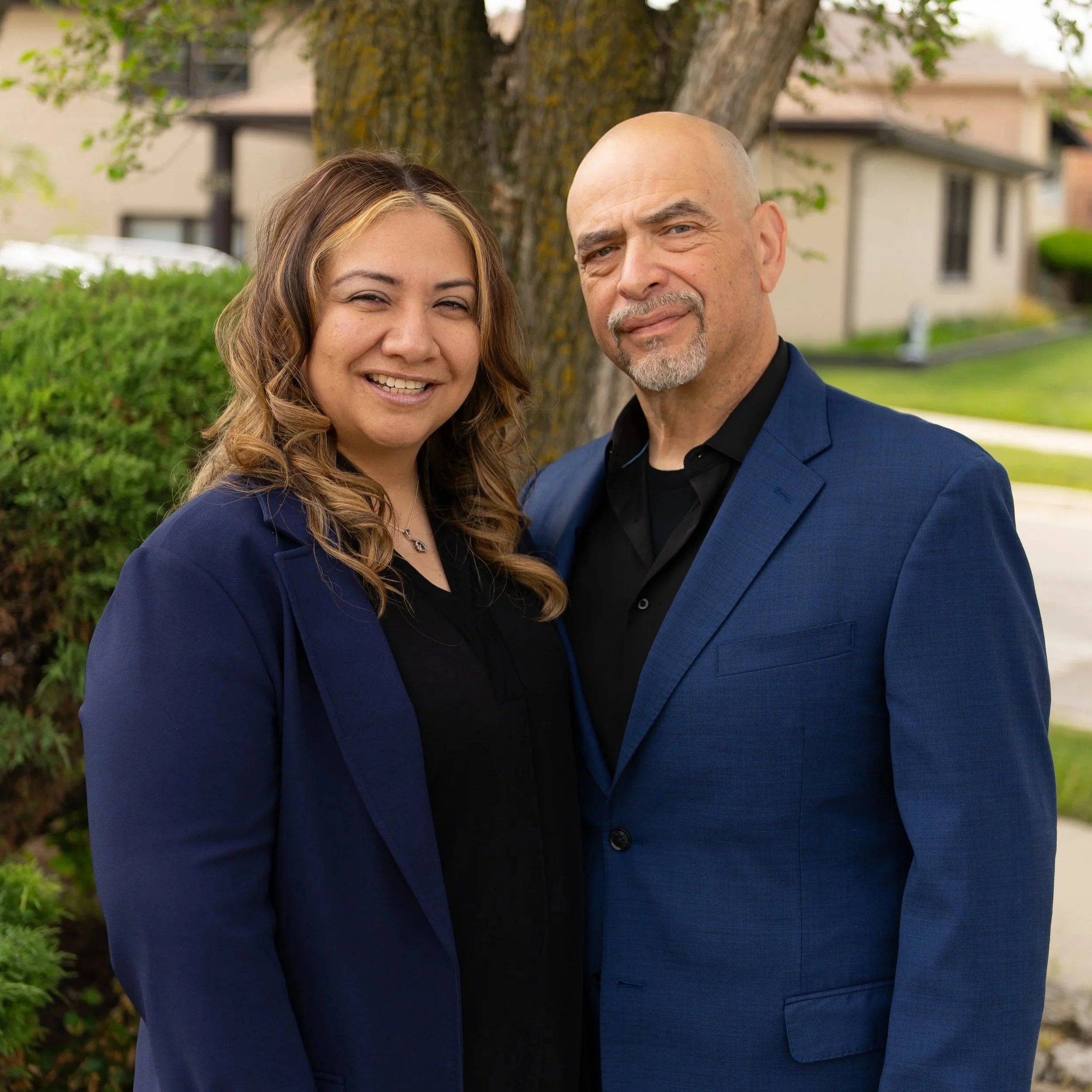 A smiling woman and a man in a blue blazer standing outdoors in front of a tree and residential buildings.