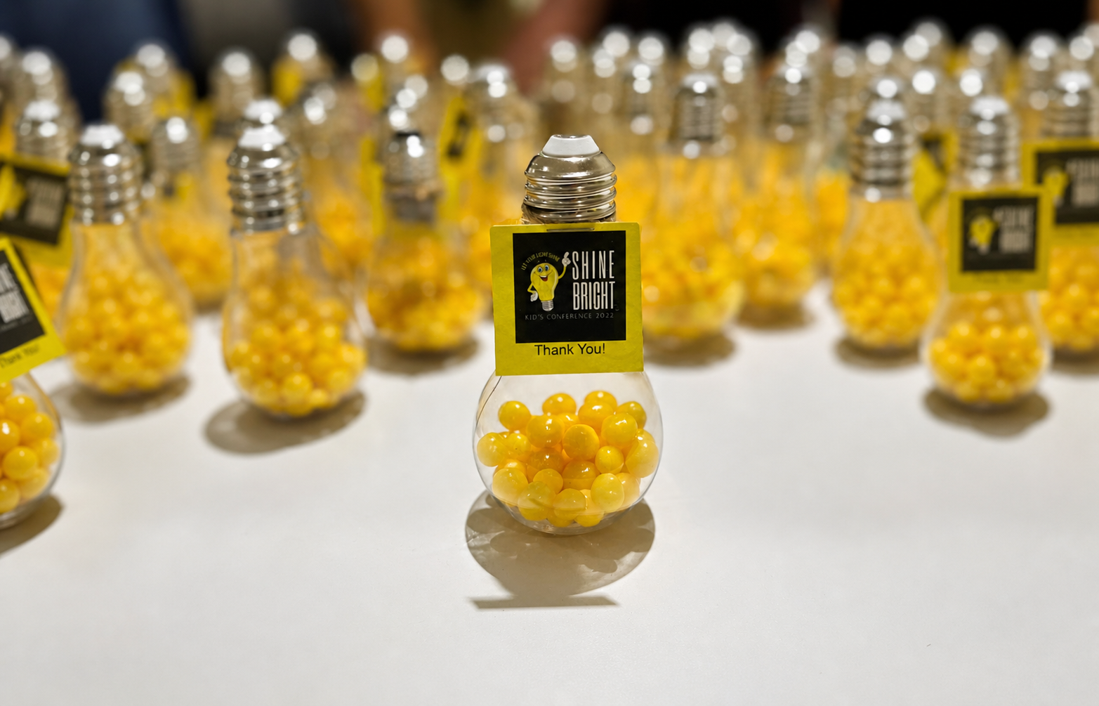 A display of small lightbulb-shaped containers filled with yellow gumballs at a conference, with one container in focus and a label that reads, "SHINE BRIGHT. Thank You!"
