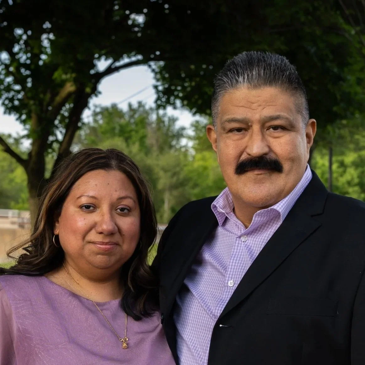 A man and woman standing outdoors, with green trees and a cloudy sky in the background. The man has a mustache, slicked-back hair, and is wearing a dark suit jacket with a light purple shirt. The woman has shoulder-length dark hair, is wearing a purple top, and has a gentle smile.