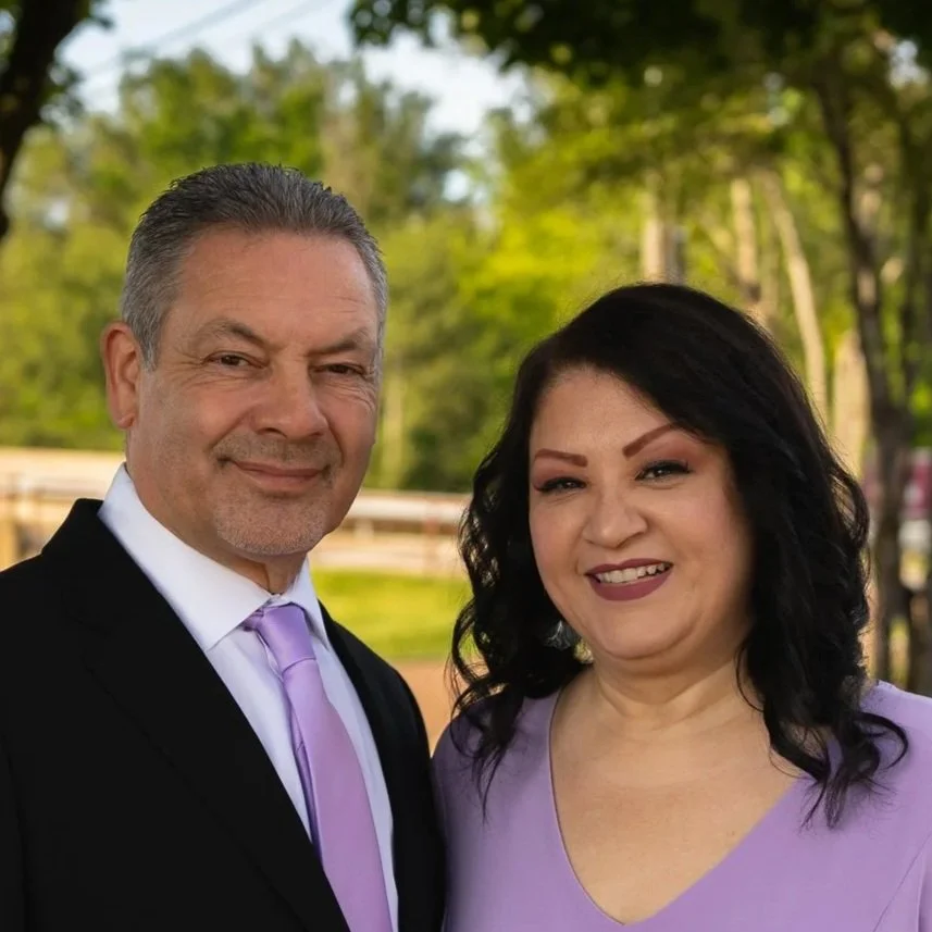 A man and woman standing outdoors, smiling, with trees in the background. The man is wearing a black suit, white shirt, and lavender tie. The woman is wearing a purple top and has black hair.