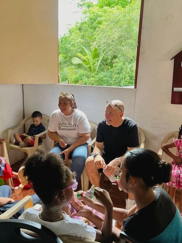 A group of children and two adults sitting together in a bright room with large window showing greenery outside, engaging in a joyful activity.