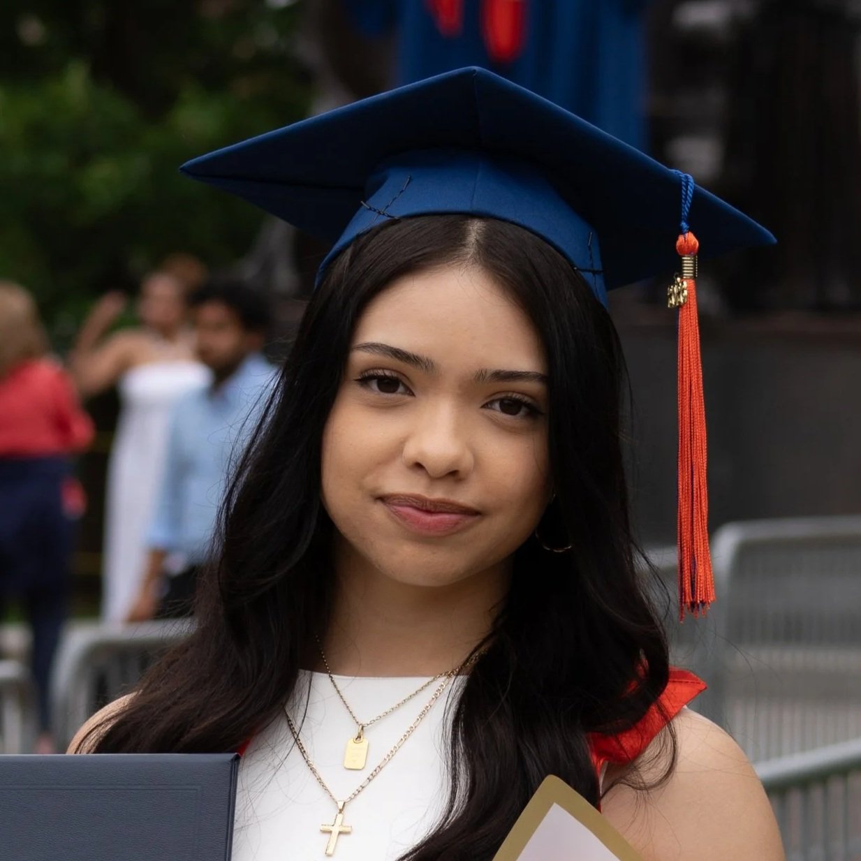 Young woman wearing a blue graduation cap with an orange tassel, holding a diploma and smiling outdoors at a graduation ceremony.